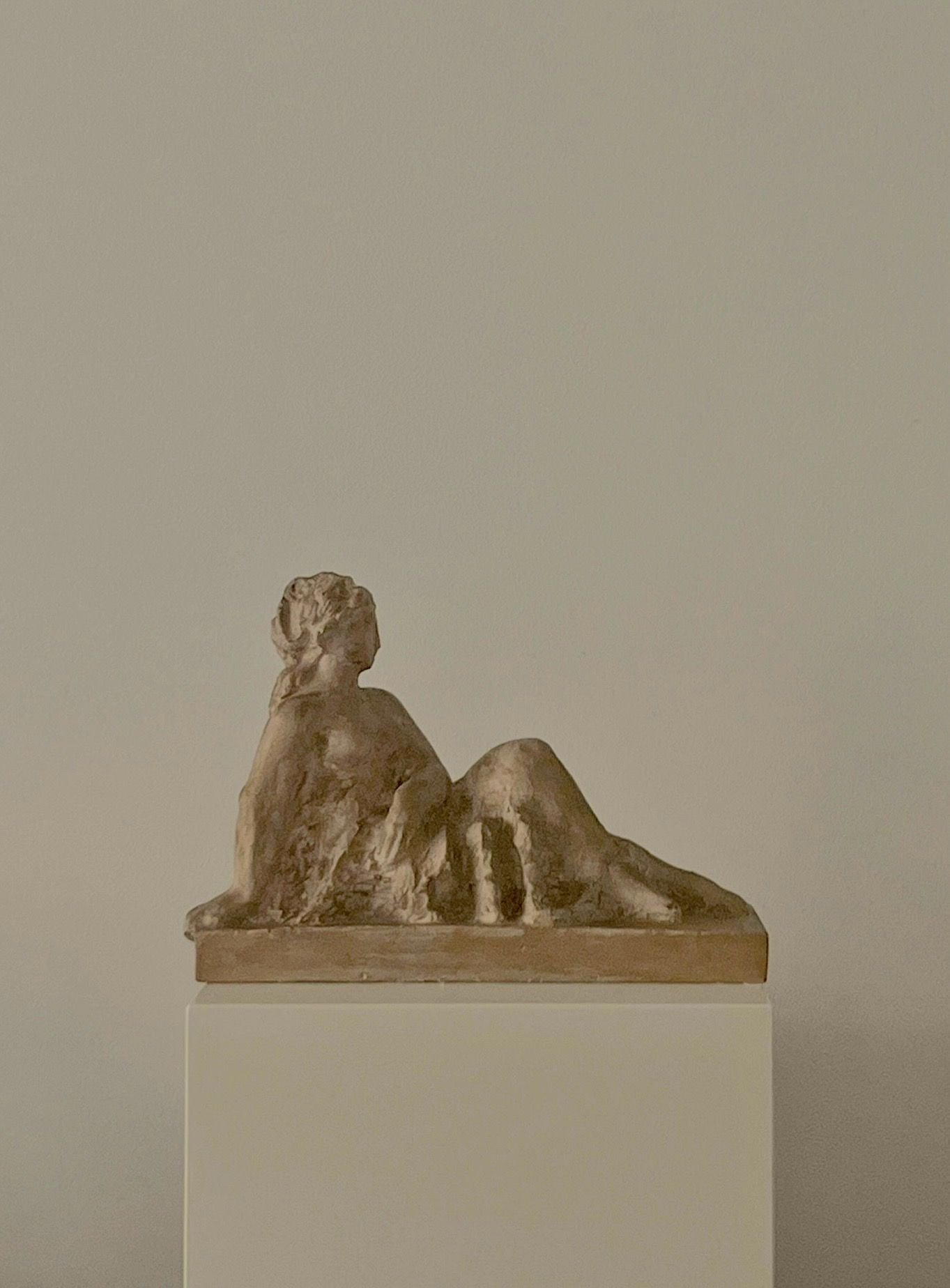 The Sculpture by Sakari Tohka 1938 from Médecine features a small bronze reclining female figure, relaxed and leaning on one arm with legs extended, displayed on a white pedestal against a light beige background.