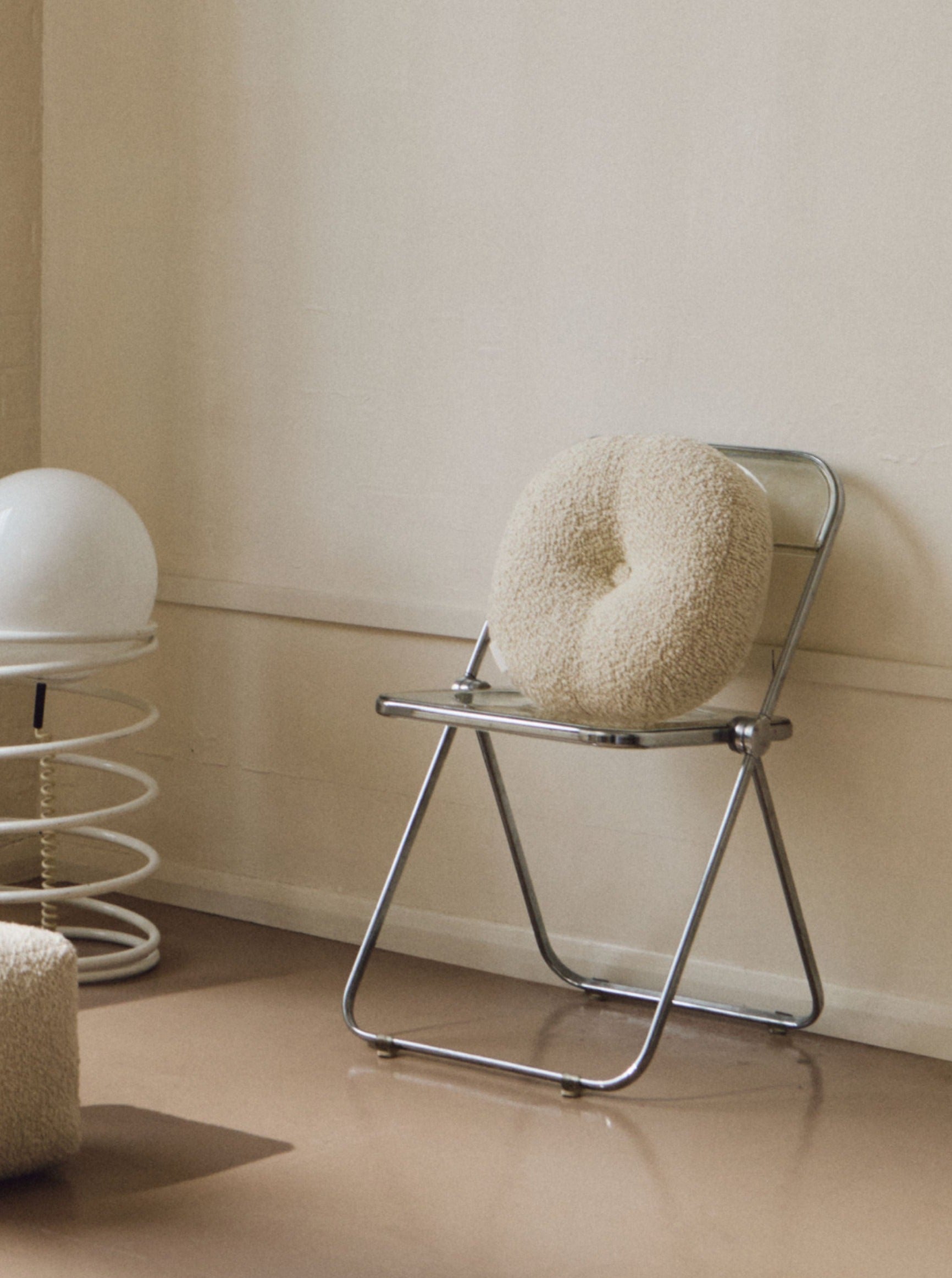 A minimalist room featuring a metal folding chair with a handmade cushion made from British eco wool, a spring-like white lamp with a round top, and Le Rond - Moyen - Wool Bouclé - Egg White by AMÉLIORÉ. The space has a neutral color palette with beige walls and a reflective, polished floor.