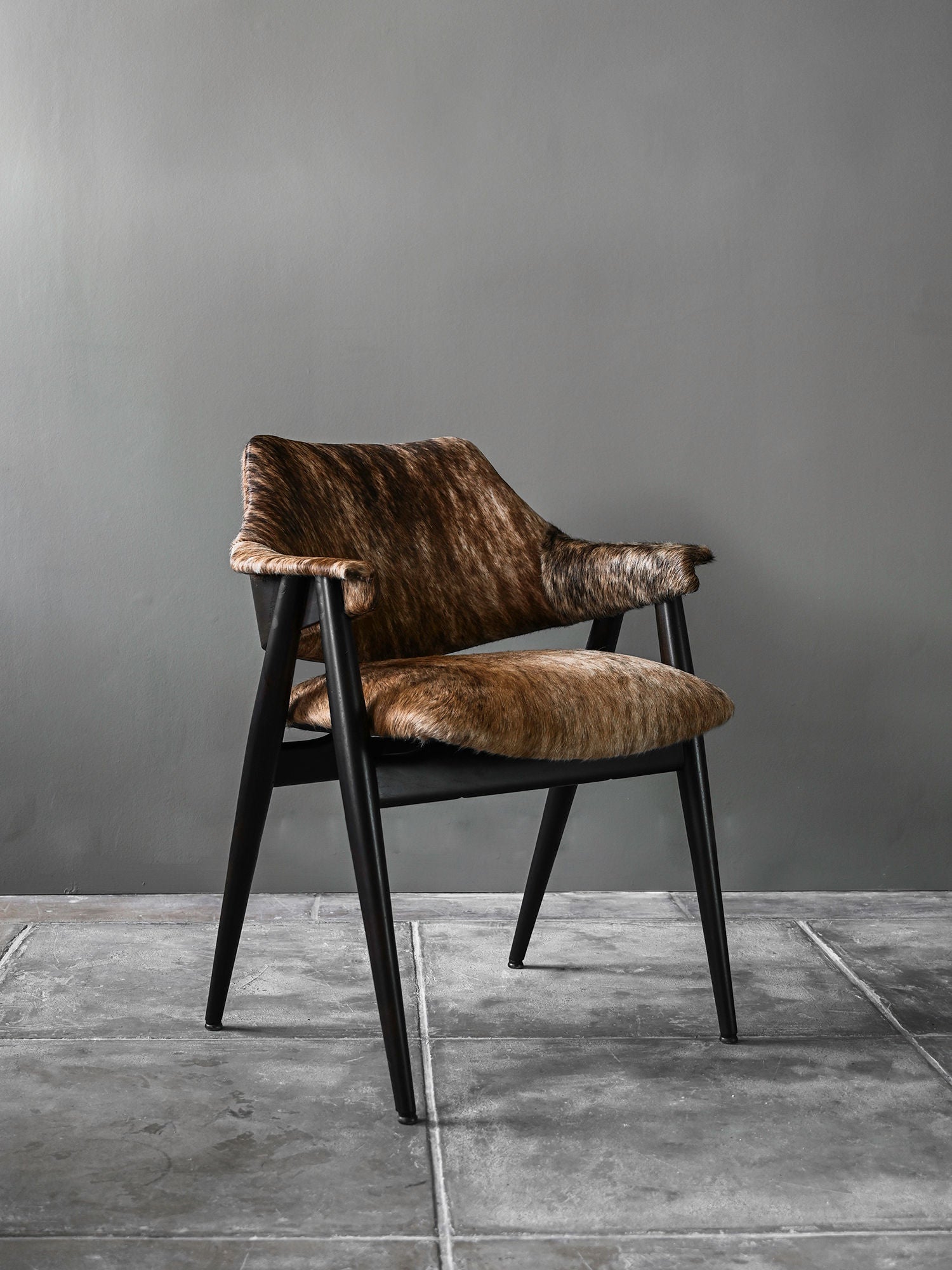 The Set of 8 Pierre Paulin Dining Chairs by Studio OSKLO features brown fur-covered seats and backrests with black wood armrests and legs, inspired by 1960s German design, shown on gray tiles against a plain gray wall.