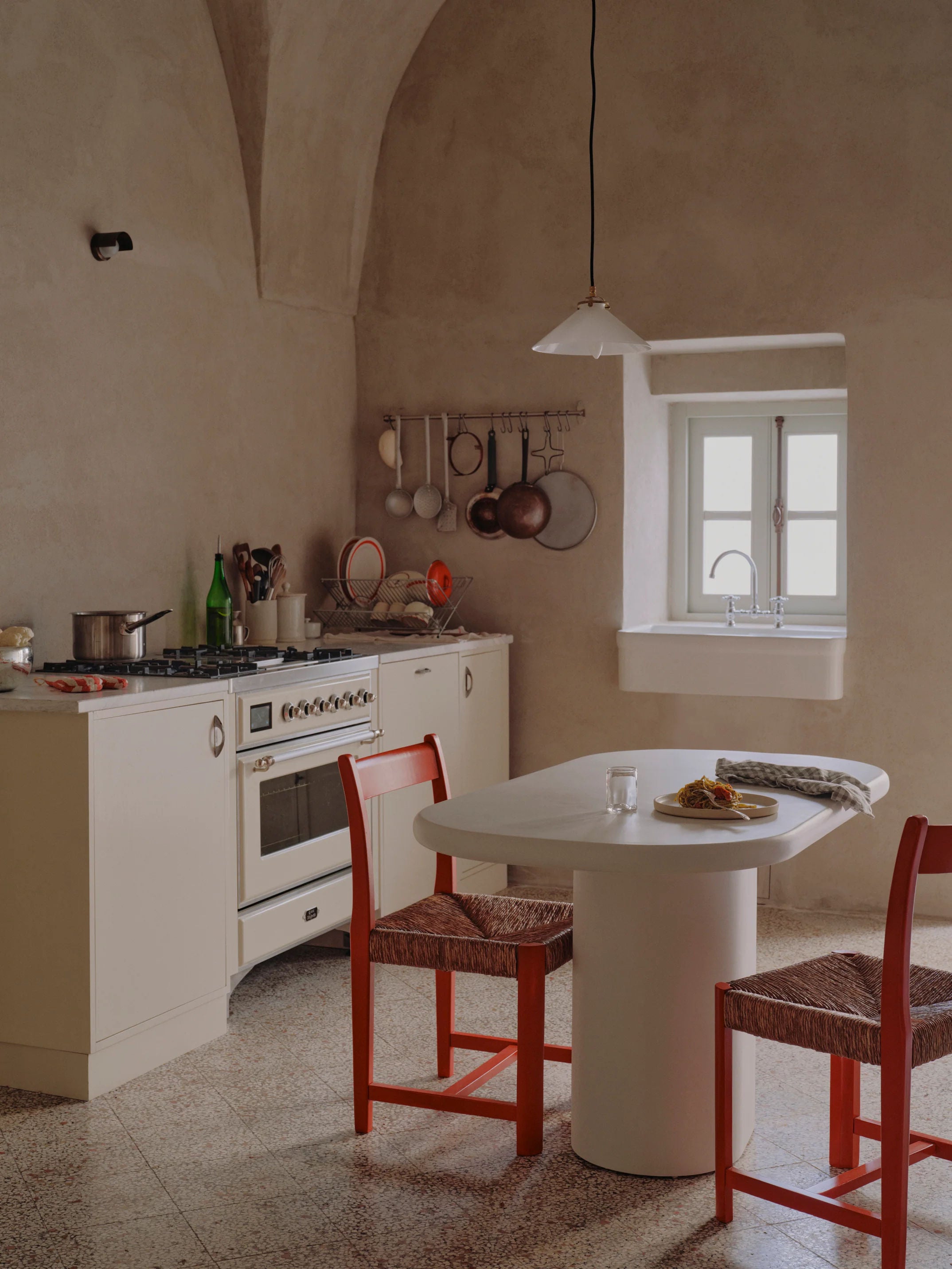 A cozy kitchen exudes rustic charm with a MarlotBaus Lucca dining table, its oval central leg flanked by two red chairs. A stove sits in the corner, with pots and utensils hanging above. Natural light pours through a small window above the micro cement sink.