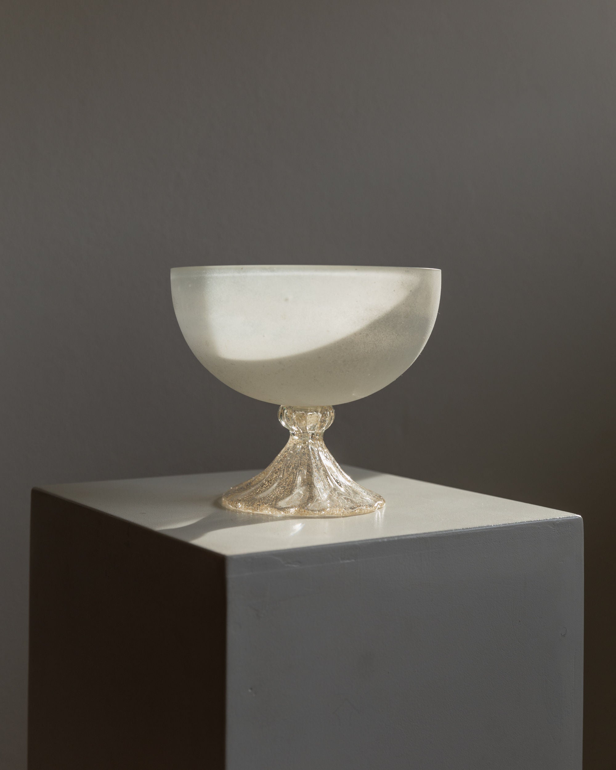 Displayed on a white pedestal, the Murano Glass Bowl - Italy 1970s by Bottega Jacobs features a decorative base. Frosted glass and its base are softly lit by natural light, casting subtle shadows against a muted gray background.