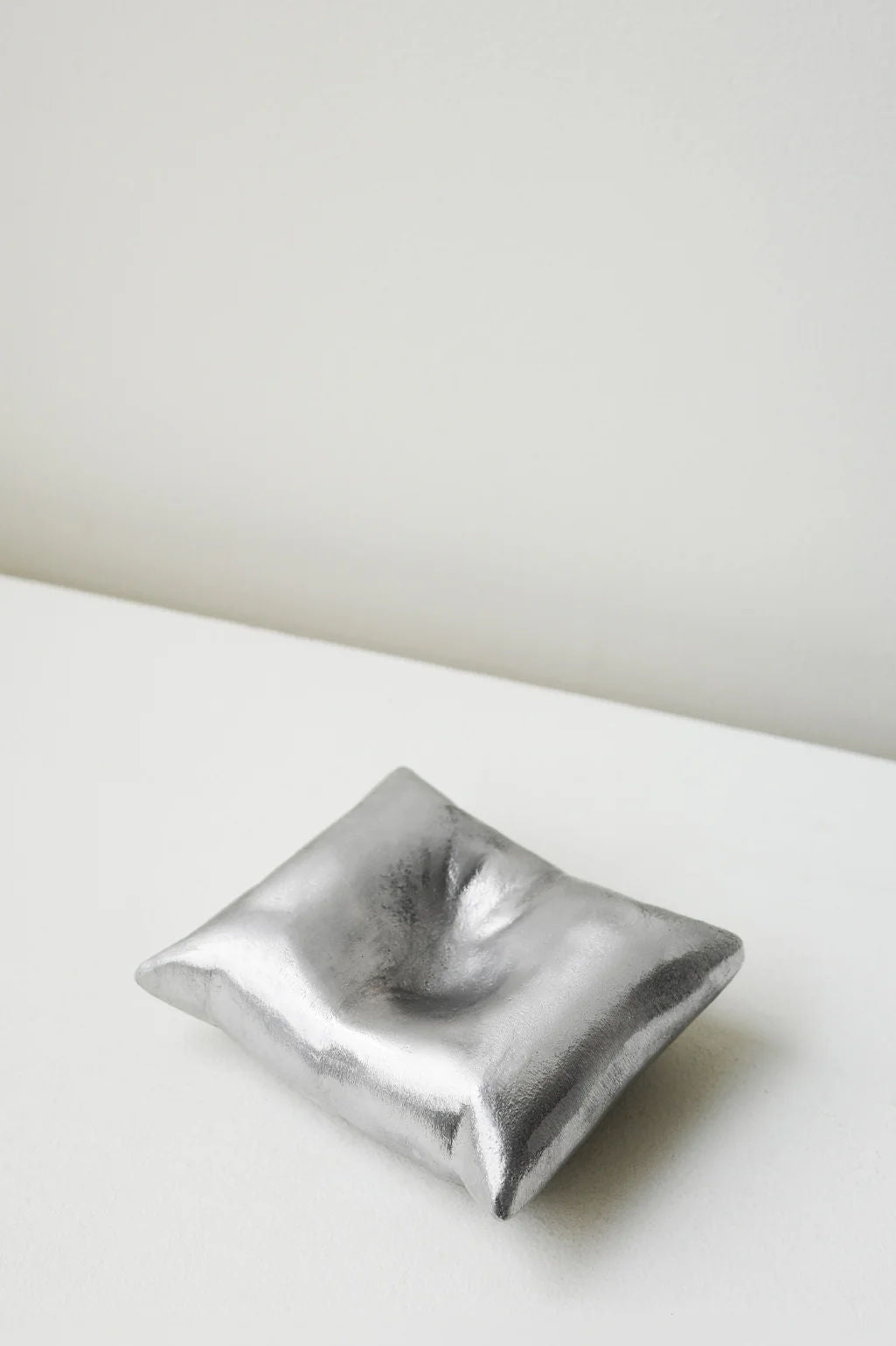 The Completedworks Bumped - Aluminium Cushion in Silver, a small rectangular pillow with a dimpled center, rests on a light surface against an off-white wall, resembling a unique handcrafted bowl.