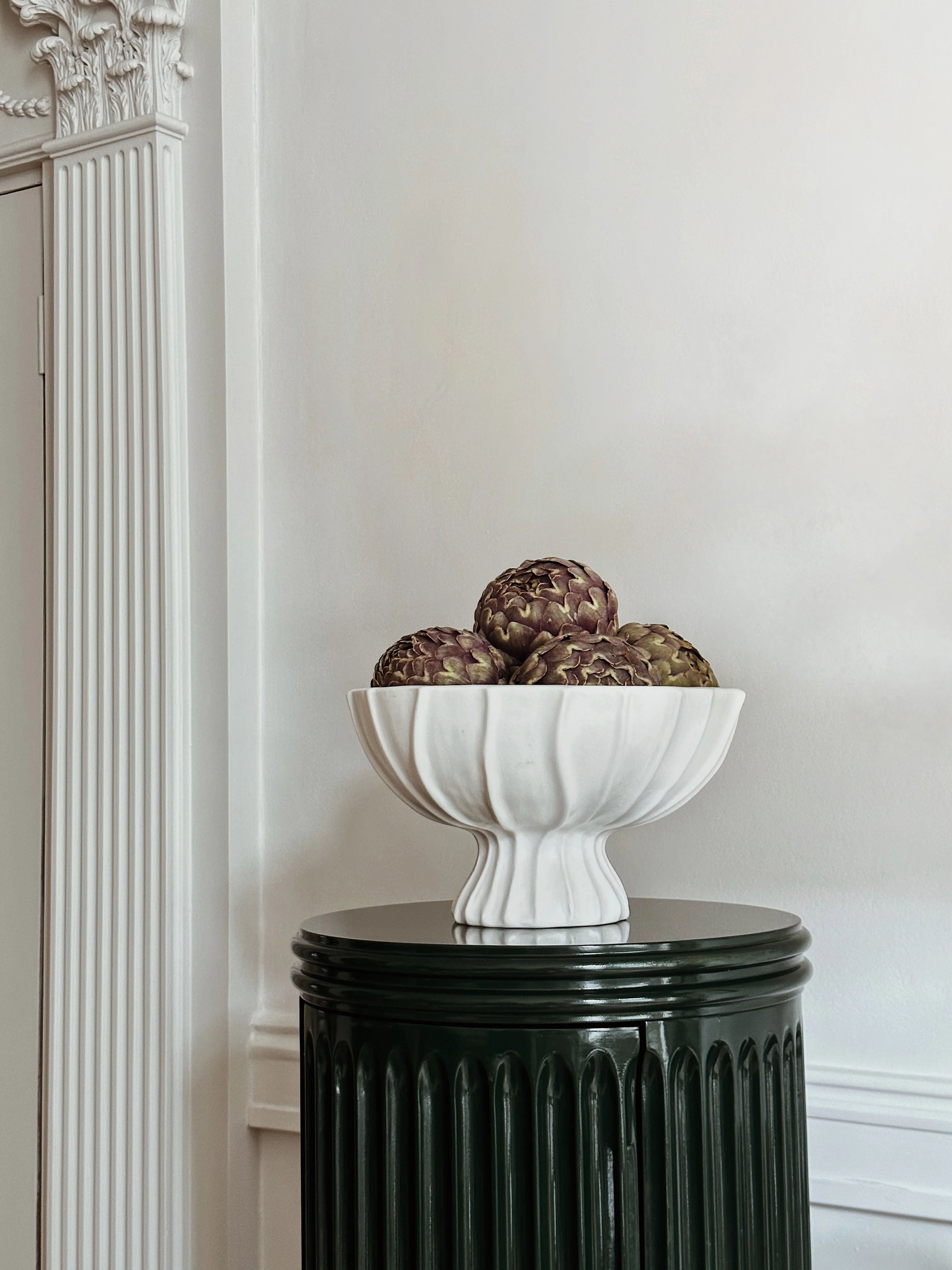 The Cloak Pedestal Bowl by Ransom & Dunn, a highlight from our handmade in Portugal collection, showcases artichokes beautifully as it sits elegantly on a dark green pedestal. The white walls and decorative molding in the background amplify the classic elegance of this refined setting.