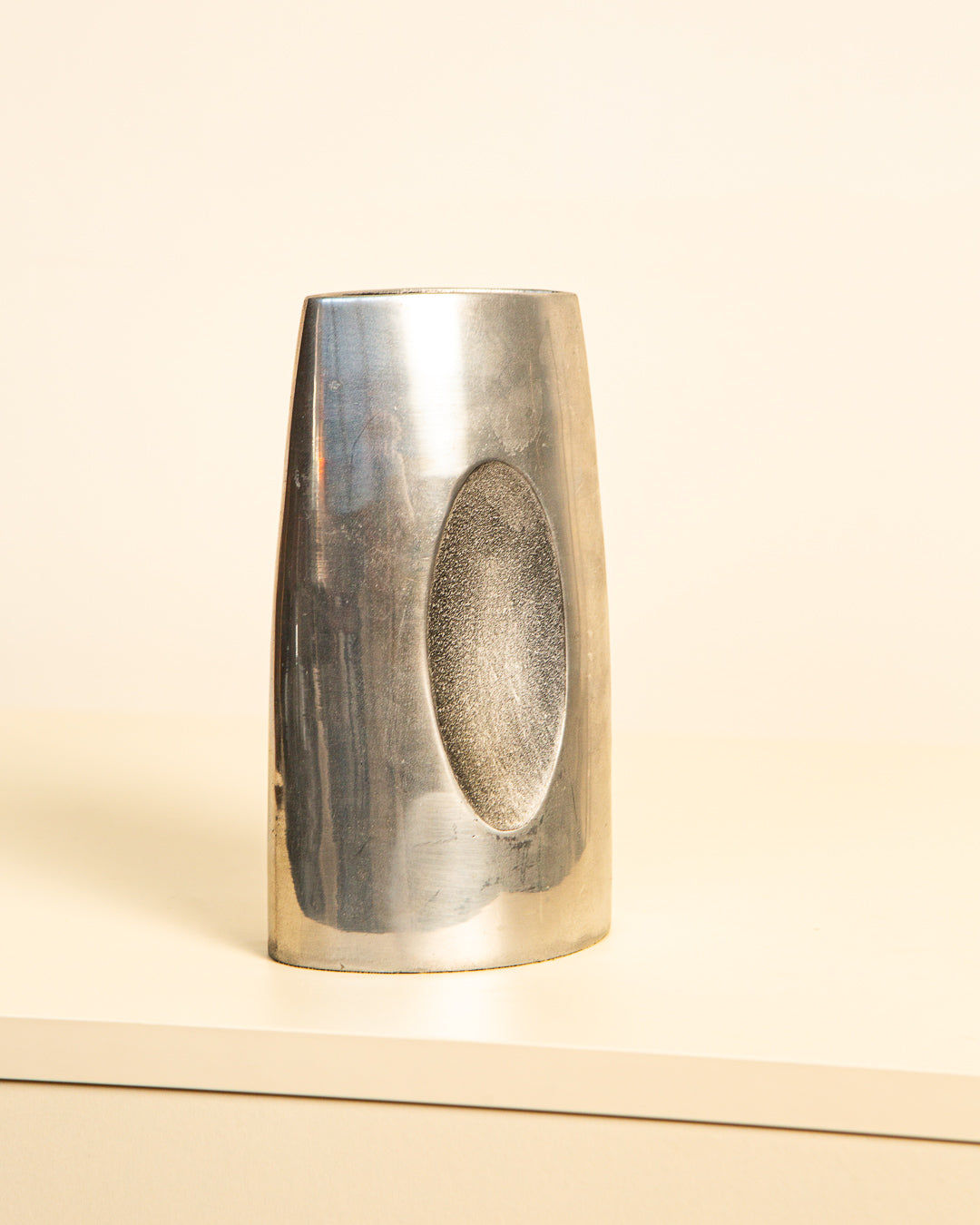 The Treaptyque Italian Vase in Aluminum 80's features a sleek, reflective surface with gently curved sides and an oval indentation. This minimalist design elegantly decorates light surfaces against neutral backgrounds, capturing subtle reflections on its polished exterior.