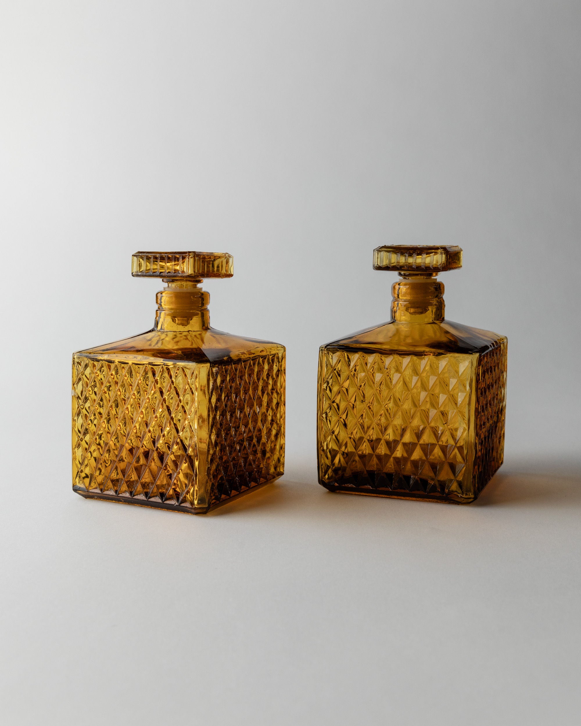 Two Bottega Jacobs Amber Glass Decanter 70s, featuring square shapes, vintage diamond point patterns, and flat round stoppers, are placed side by side on a plain, light background.