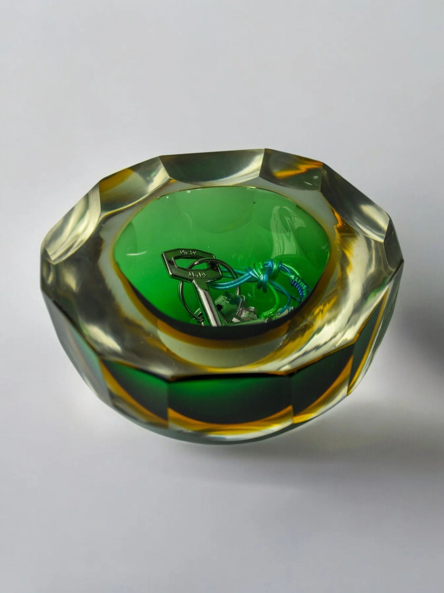A faceted green-centered Sommerso Vide-Poches bowl by Seguso from Malamar Studio cradles a shiny key and blue cord. Its transparent design mixes green and amber hues like a Murano ashtray, allowing the items to shimmer subtly through its exquisite glasswork.