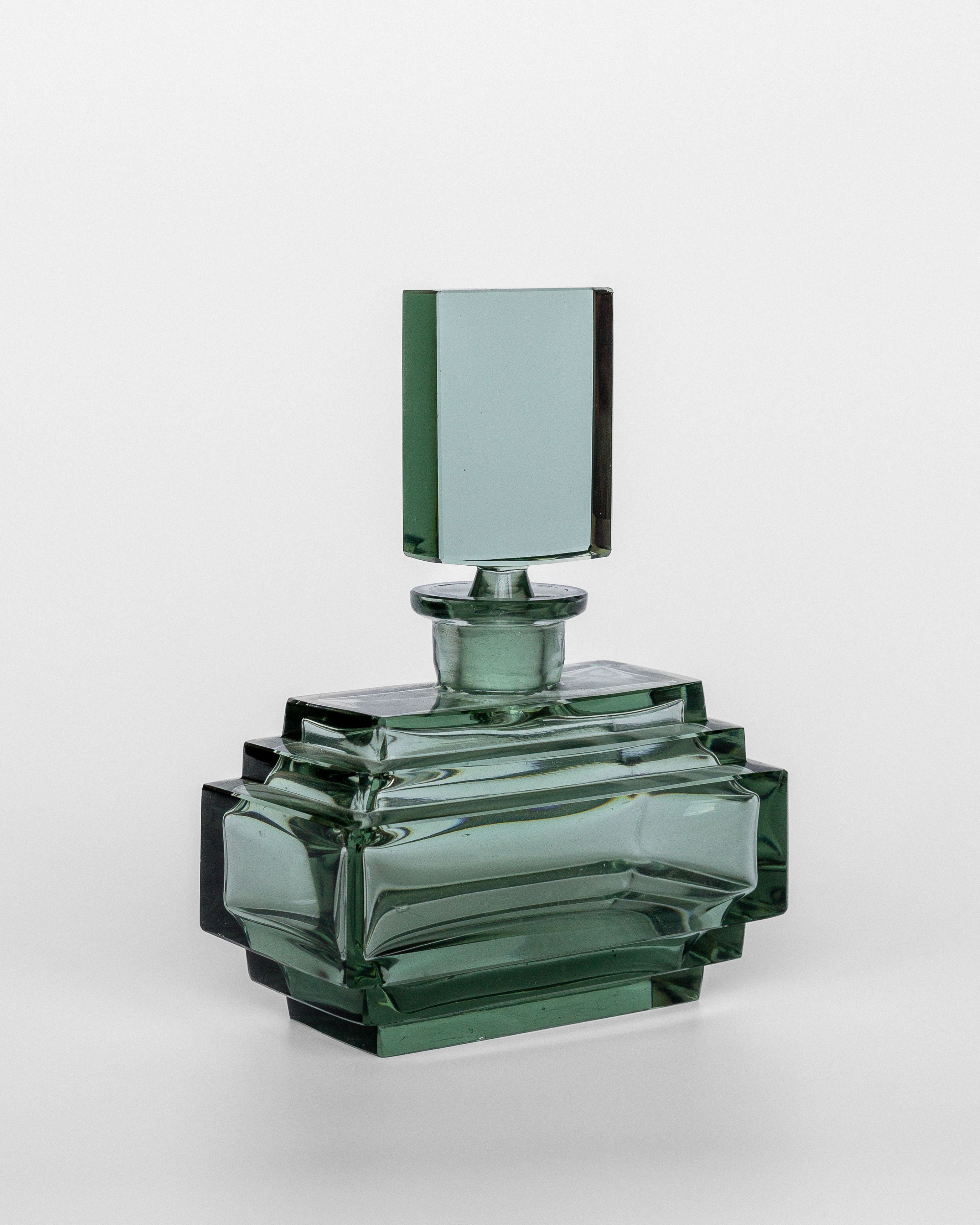 A green-tinted, geometric glass perfume bottle from Spigel, called the Art Deco Perfume Bottle, 1930s, features a rectangular stopper and sits on a plain, light background. This perfume bottle of Czech origin boasts an angular, modern design with clean lines and clear edges reminiscent of the iconic 1930s style.