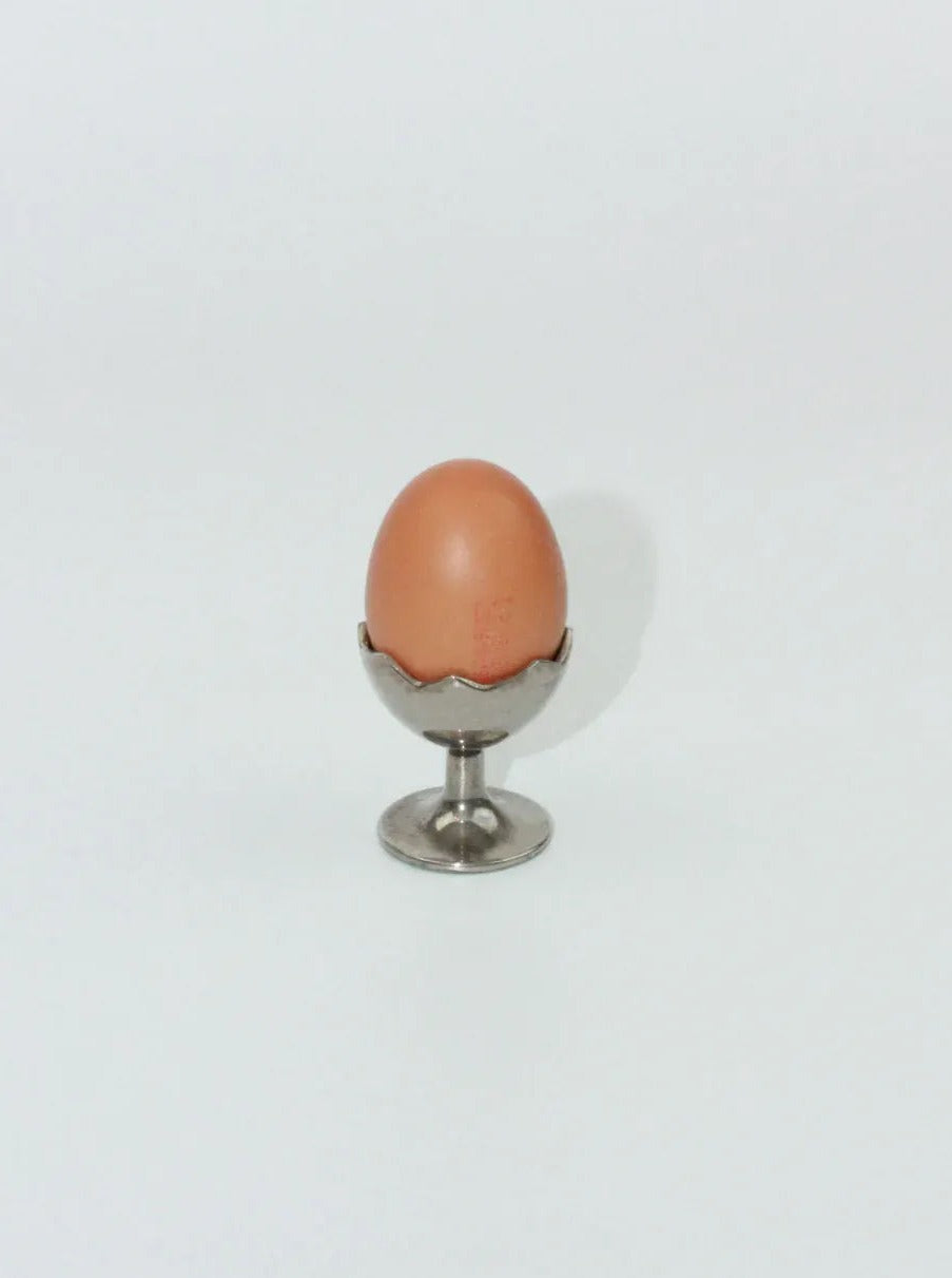 A single brown egg sits upright in a small, silver Boga Avante Shop Egg Cup with a decorative edge against a plain white background. The shadow of the egg and cup is visible to the right.