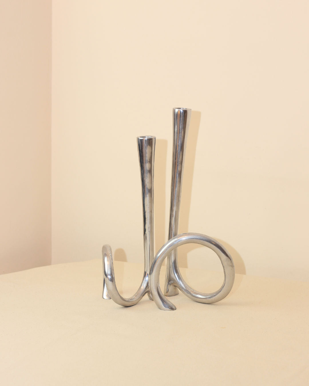 The Boga Avante Shop Cast Aluminum Candleholders feature polished silver metal with two tall, curved stems and an abstract looping base—evoking a modern 1980s candelabra—ideal for minimalist decor on any neutral surface.