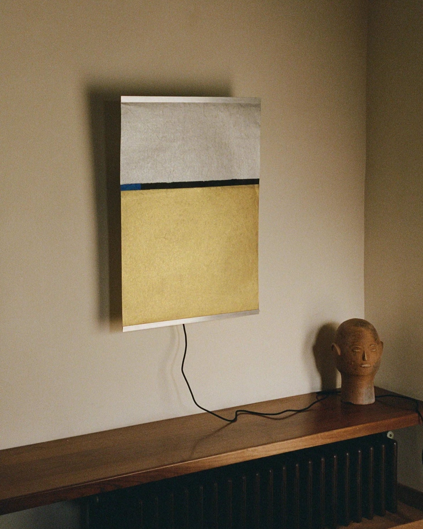 A minimalist artwork featuring sections of white, black, and yellow is mounted on a wall. Positioned below it, a wooden surface displays a handmade stone head sculpture. A cord extends from the TEGET Panel Wall Lamp – Wheat Field attached to the artwork, indicating it might be illuminated by this source.