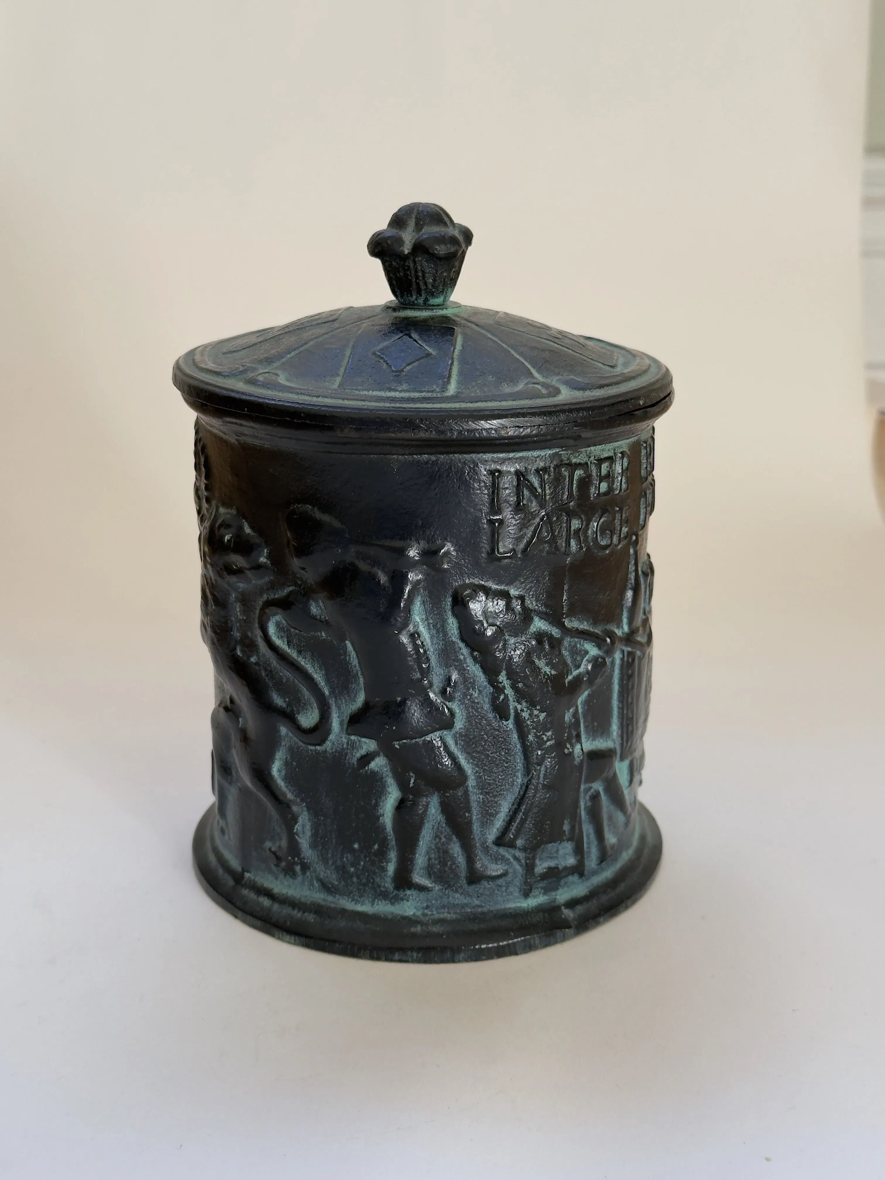 The Antique Cast Iron Tobacco Jar II by Galerie Storm is a 20th-century design featuring a cylindrical cast iron jar with raised figures, including a man and animals. Its decorative finial lid and aged metal offer historical charm, ideal for vintage enthusiasts.
