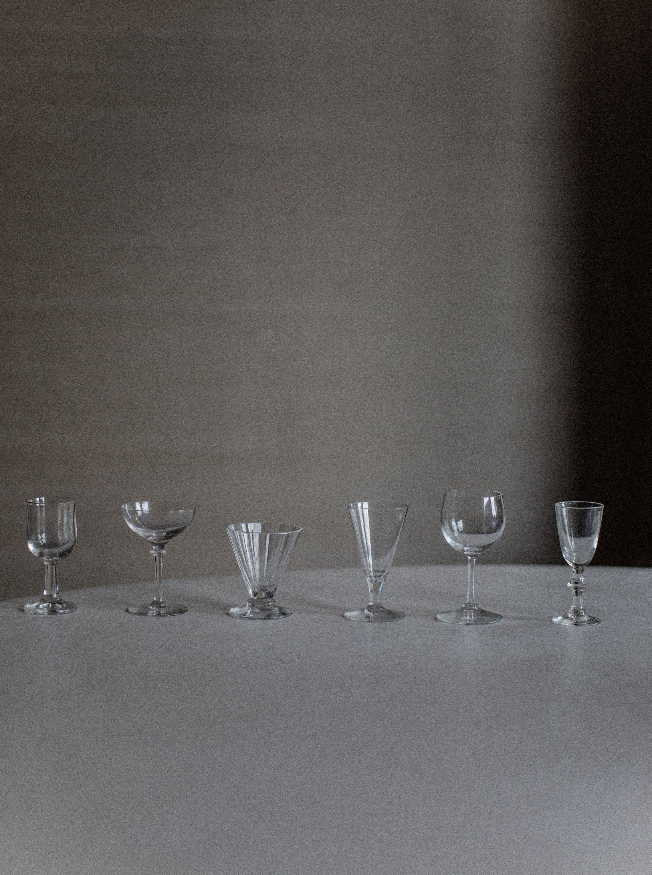 A row of six empty glasses from the "Set of Six Mix-Matched Glasses" by Out For Lunch, including wine glasses, goblets, and cocktail glasses, arranged on a plain surface against a neutral grayish background. The subtle lighting casts shadows that enhance the sophistication and create a serene atmosphere. Each glass narrates its own unique story.