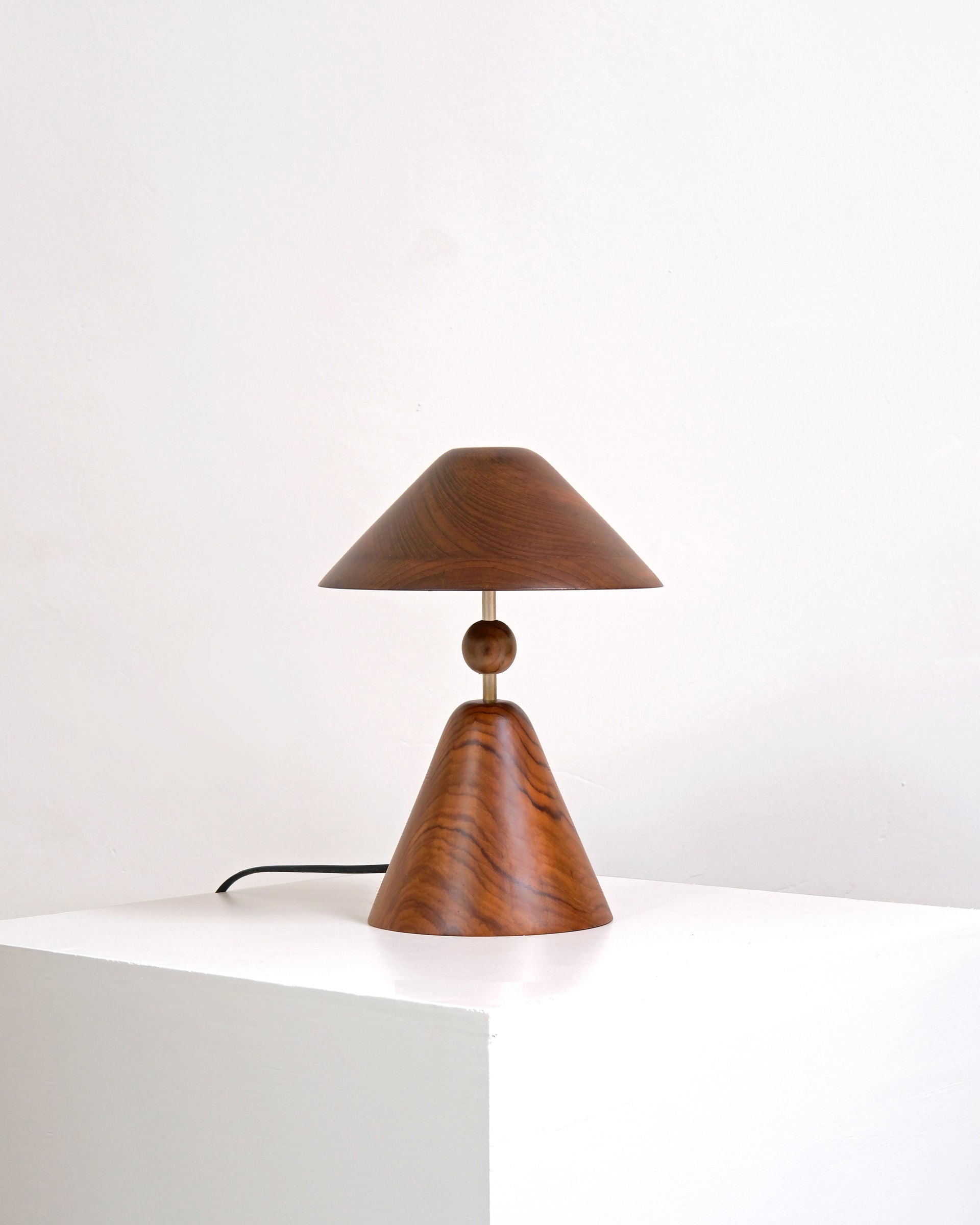 The Teak Wood Table Lamp by Studio Indigene, featuring geometric shapes and a central sphere, rests on a white pedestal. Its sculptural teak wood base and shade stand out against a plain white background.