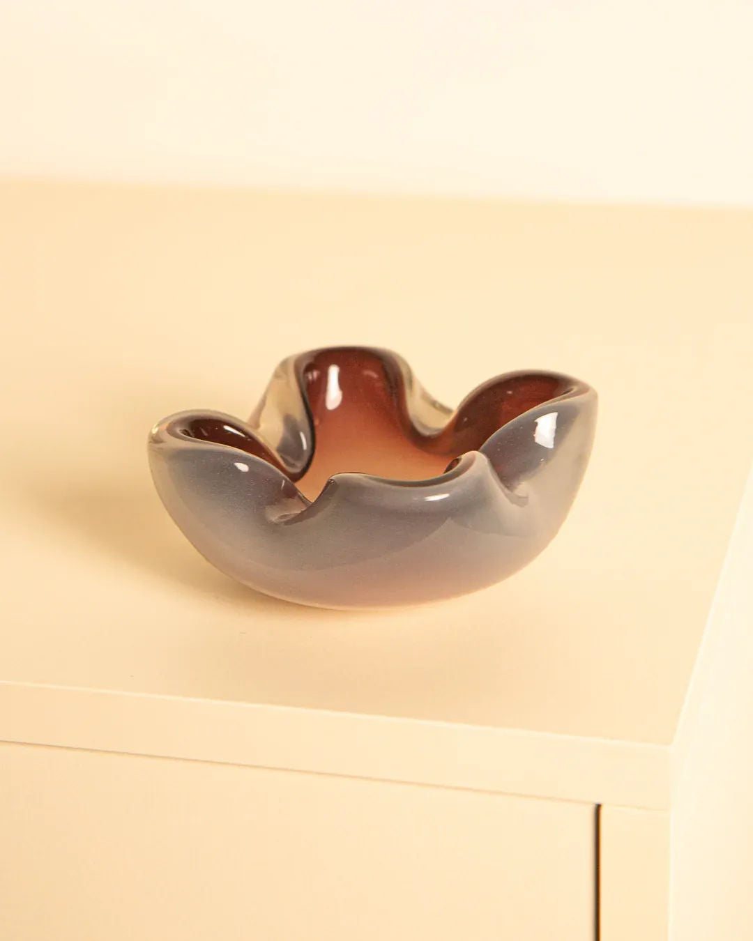 A Murano Opaque Glass "Fleur" 60s bowl by Treaptyque with a wavy edge sits on a beige surface. It features a vintage Italian-inspired gradient, transitioning from dark gray at the top to warm brown at the bottom.