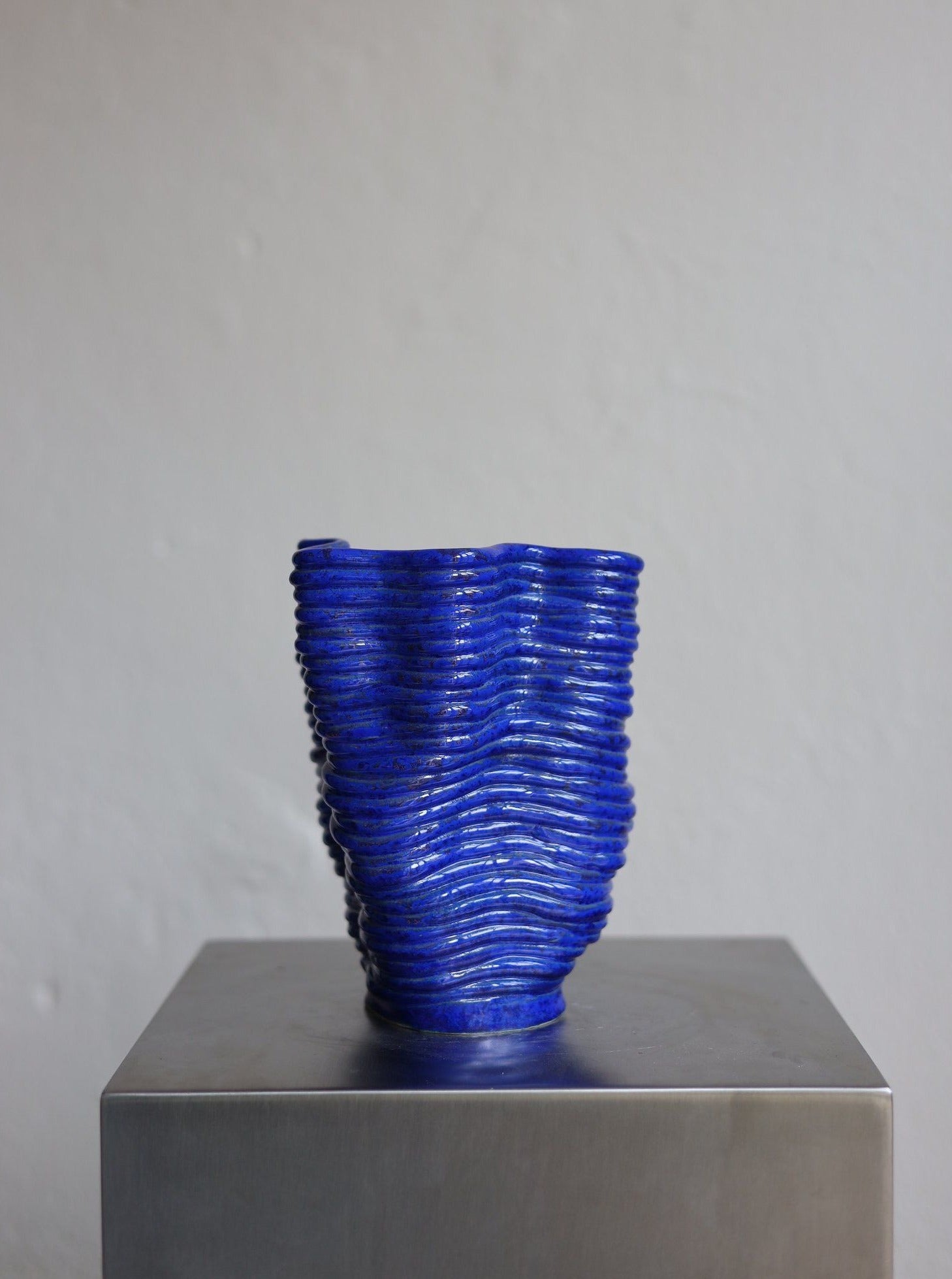 A deep blue, handsculpted Wobbly Vase Blue ceramic vase with a ribbed design displayed on a grey pedestal against a neutral wall background. (Brand Name: 2222Studio)