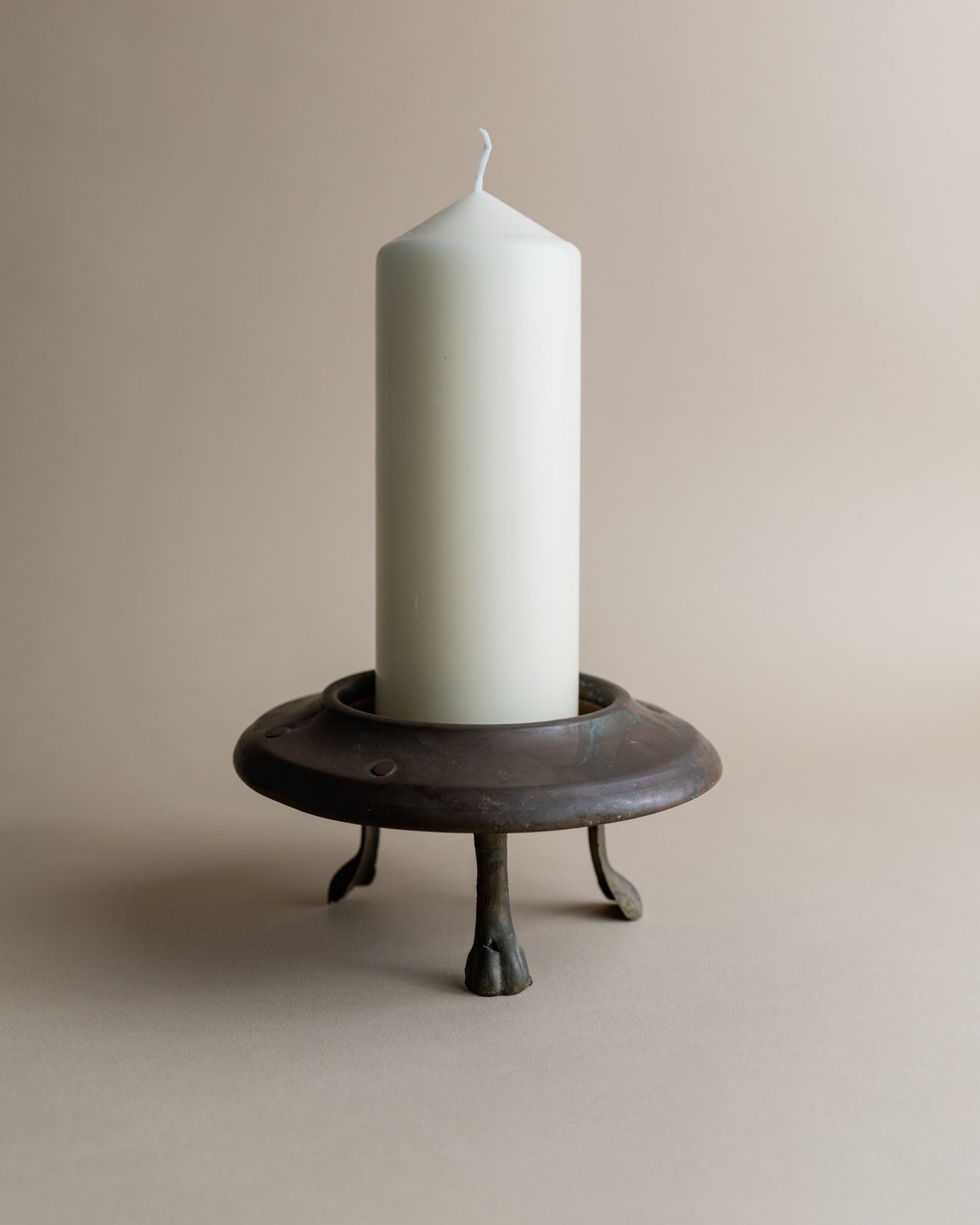 A tall white pillar candle sits on a Bottega Jacobs Vintage Copper Candle Holder with three small legs, set against a plain beige background.