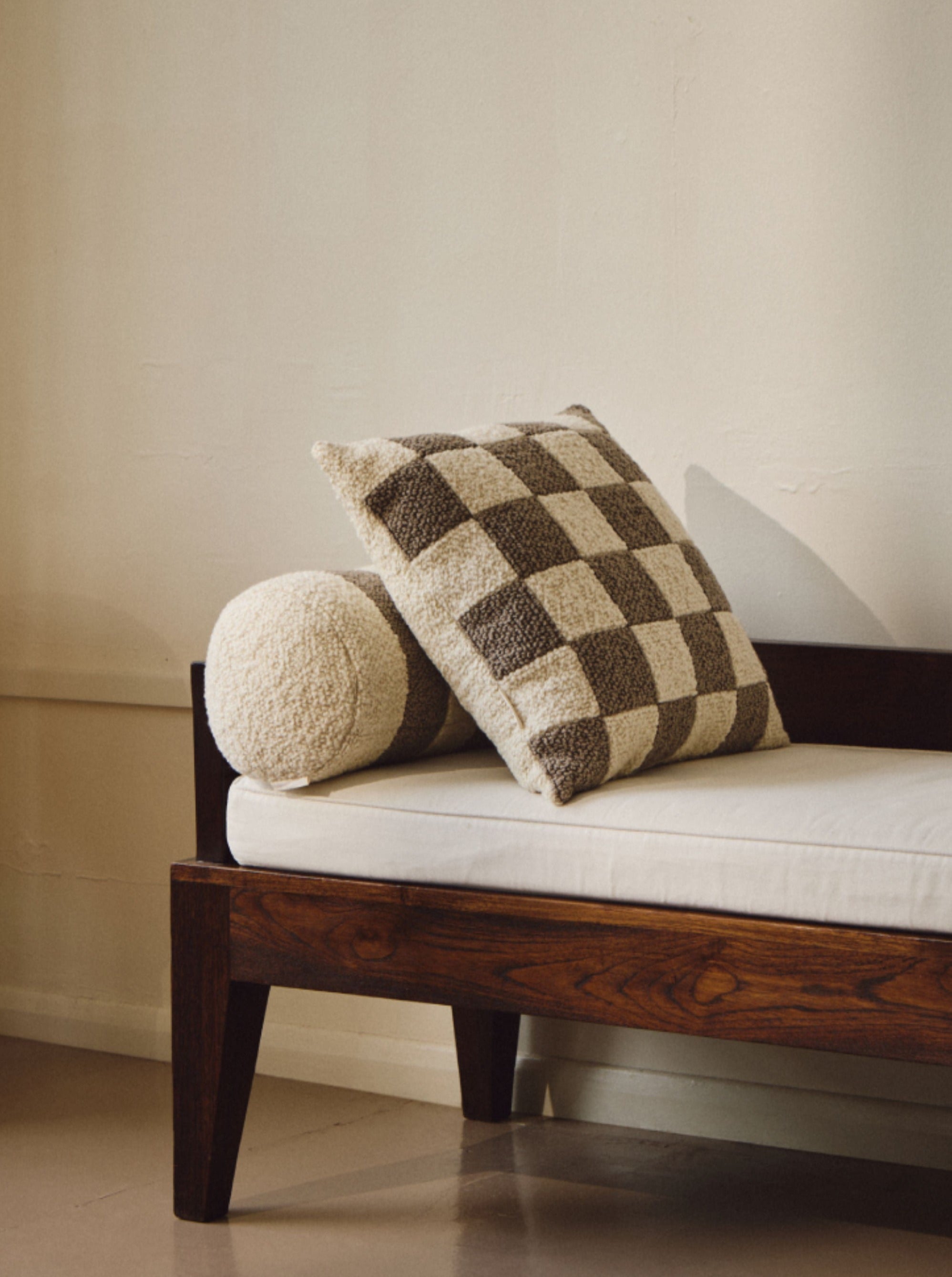 A wooden bench features a white cushion seat with a handmade cushion, including a rectangular beige pillow and a square plush checkered pillow in brown and white leaning against the backrest. The vintage-inspired design is complemented by the cream-colored wall behind and the light brown floor. Adding to its elegance is AMÉLIORÉ's Le Carré - Wool Bouclé - Sawdust/Eggshell throw blanket draped gracefully over one side.
