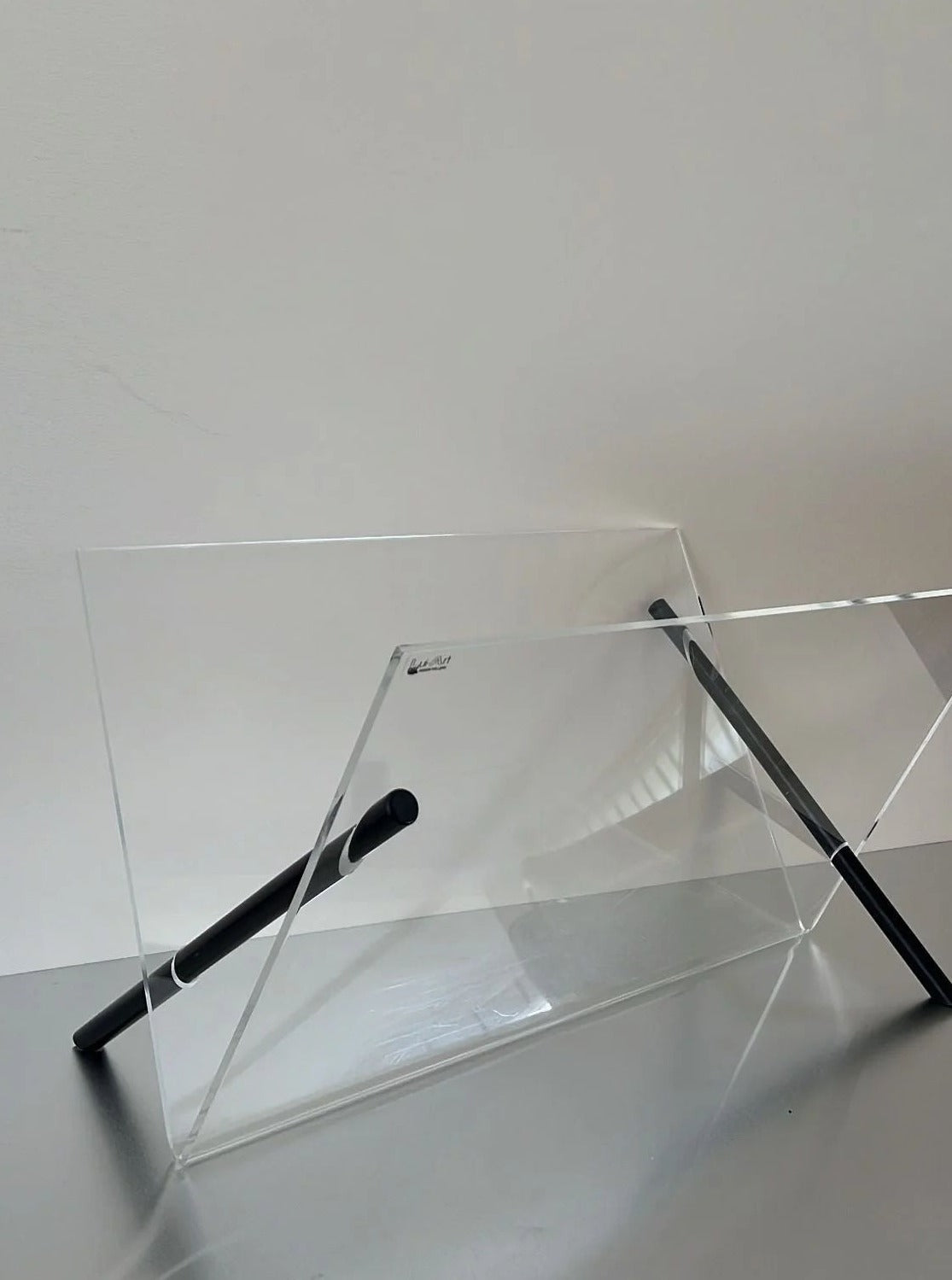 A minimalist, transparent premium plexiglass Magazine Holder Plexiglass by Lui Art Design leans against a plain wall. Two black cylindrical rods intersect with the magazine holder at a diagonal angle, possibly serving as supports. The scene is simple and uncluttered, emphasizing geometric shapes and light reflections, embodying CU-RA-TED elegance.