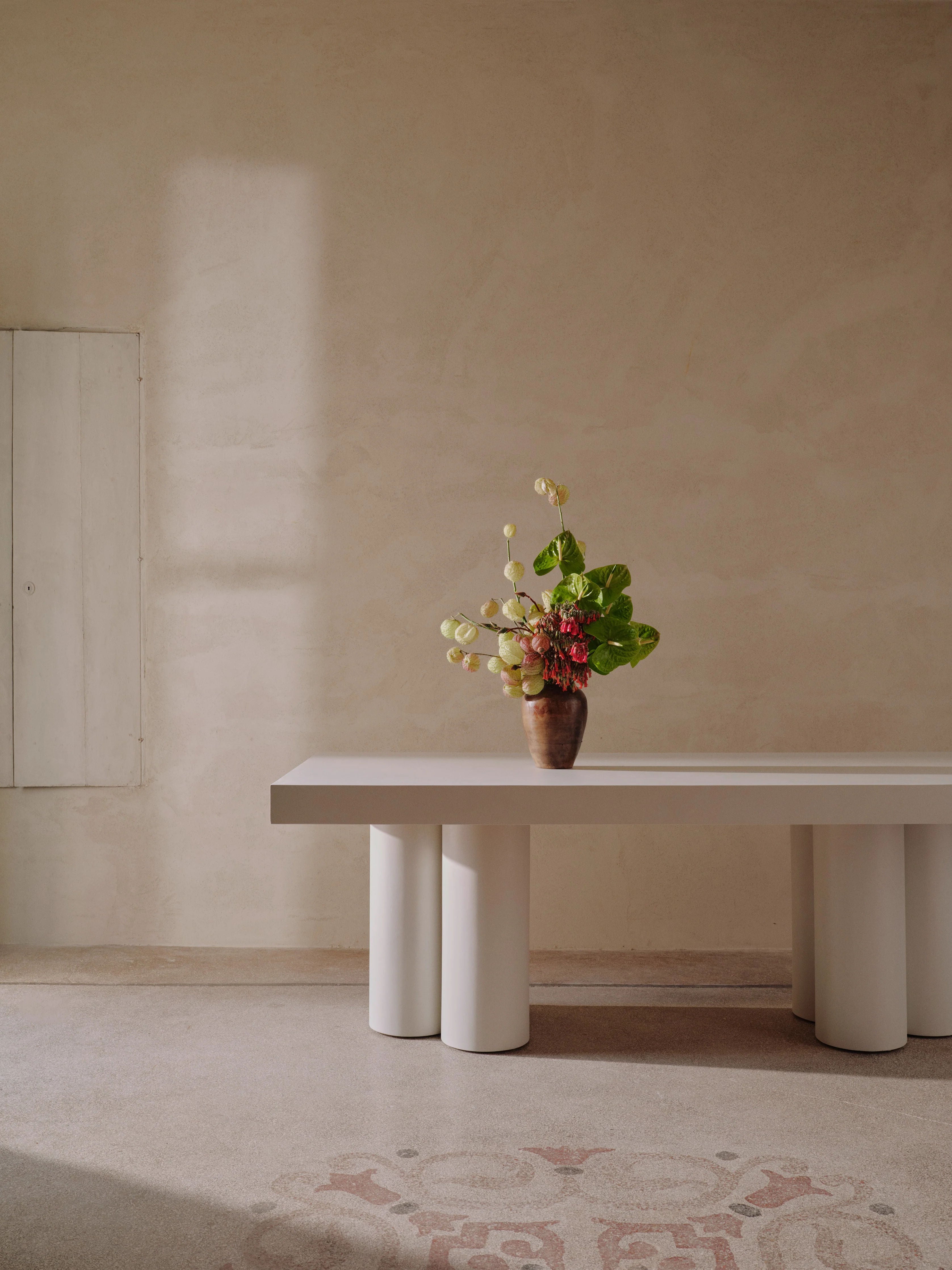 A minimalist interior features the "Bellini" Dining Table by MarlotBaus, with cylindrical legs showcasing traditional craftsmanship. The table is adorned with a vase of vibrant flowers, including green and red blooms. Soft beige walls and a patterned floor enhance the serene, elegant atmosphere as natural light creates gentle shadows.