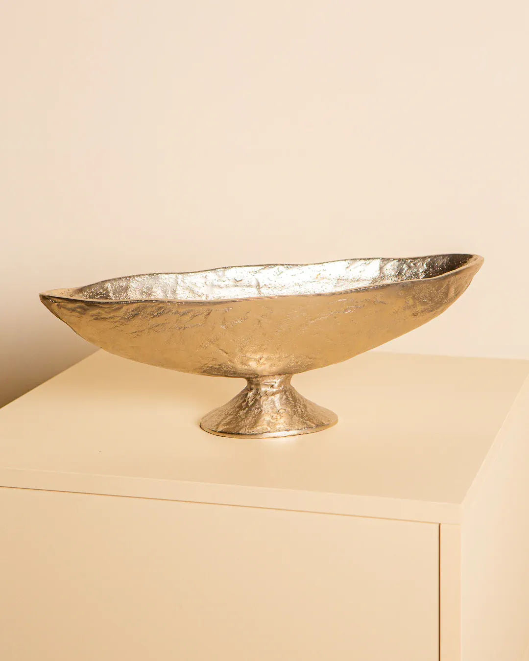 The Treaptyque Large Italian Bowl in Cast Aluminium 80's, oval and gold with a textured surface, sits on a square off-white pedestal. This Milan-inspired piece is large and shallow with a small round base against a plain, pale wall.