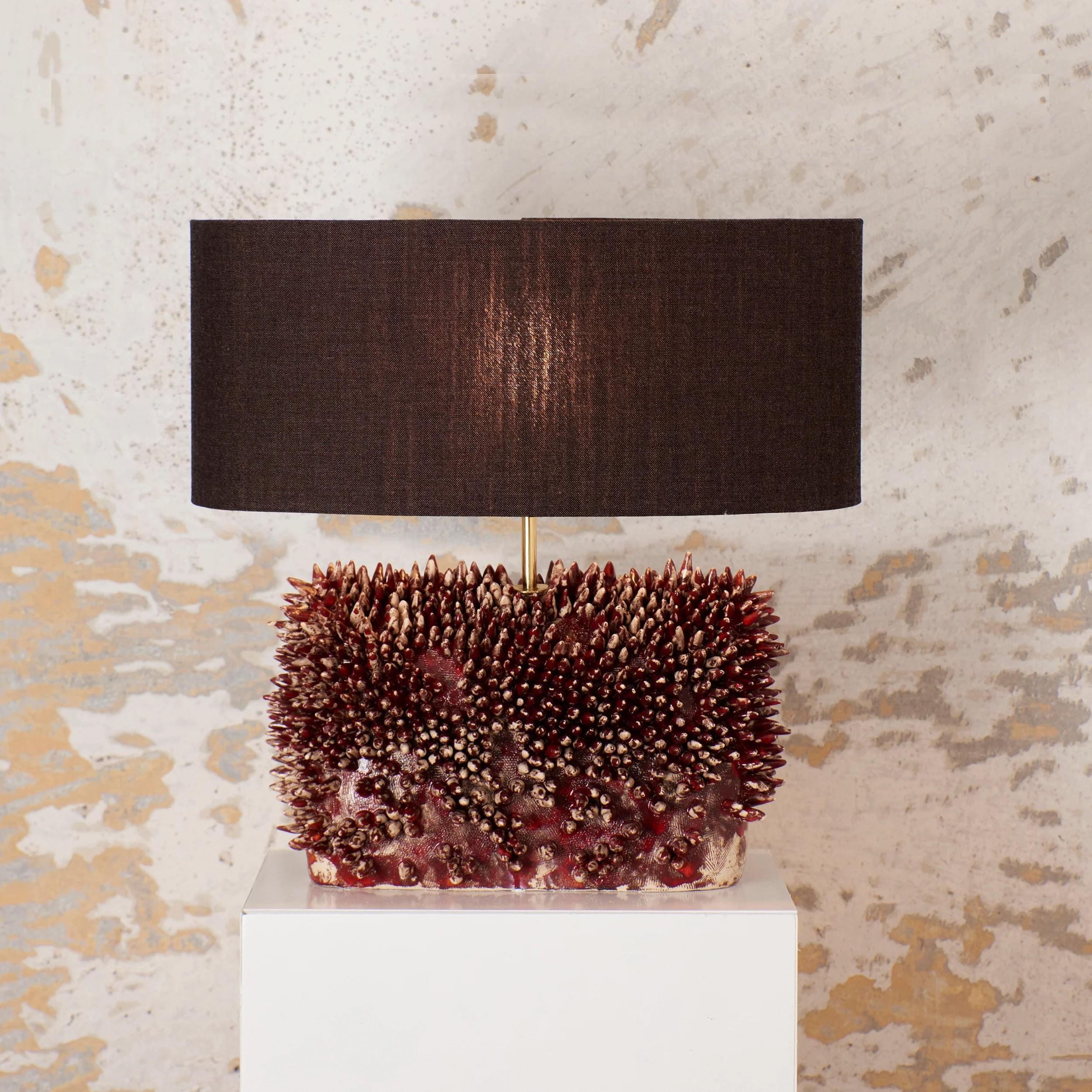 The Appuntito Ceramic Lamp by Project 213A features a dark brown cylindrical shade on a white pedestal, showcasing a minimalist design. Its coral-inspired red and brown spiky base complements its setting against a distressed beige wall—ideal for luxury interiors.
