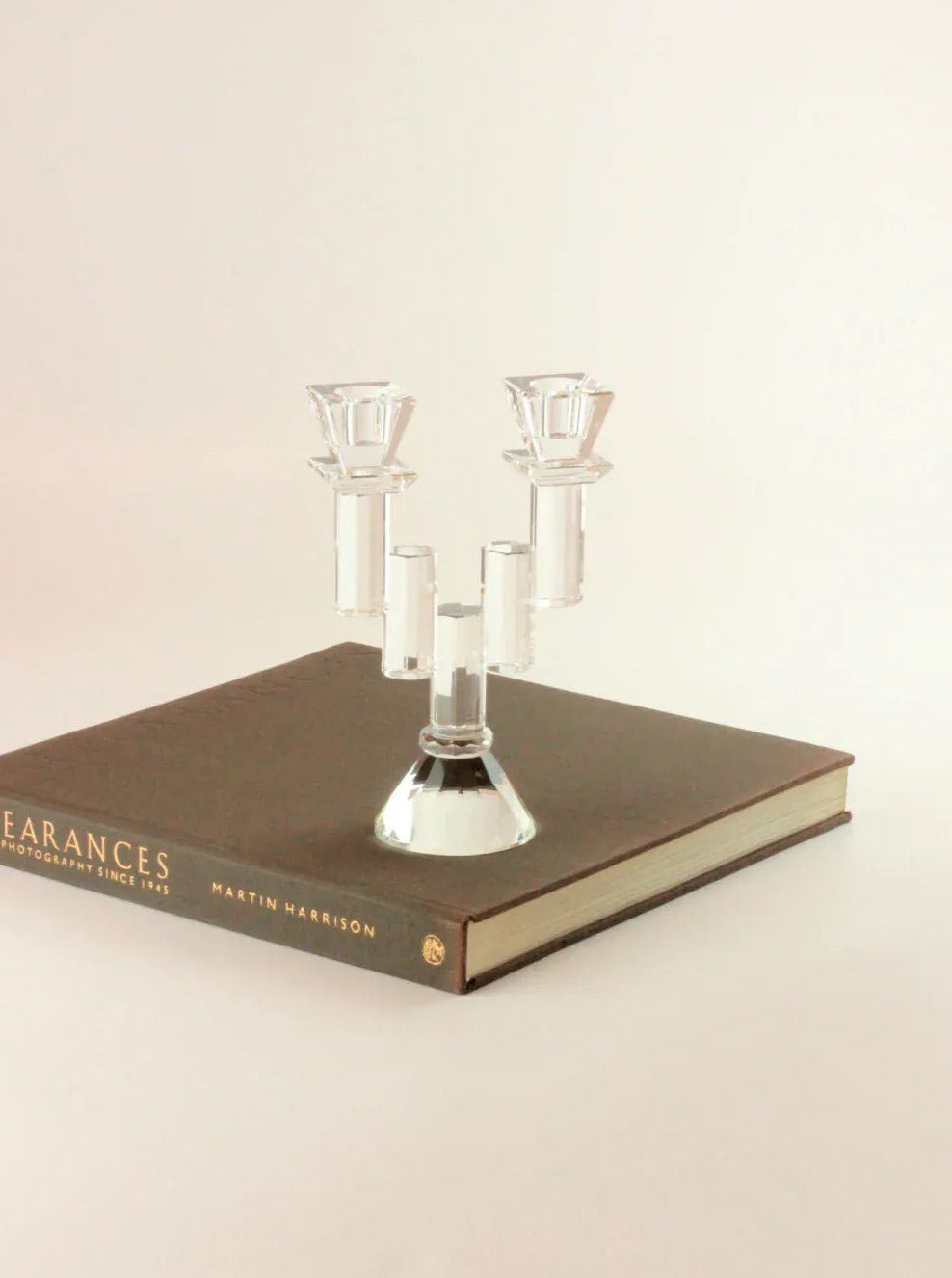 A Faceted Crystal Candleholder by Boga Avante Shop, evoking a vintage item, is placed on top of a closed hardcover book titled "Appearances: Photographs Since 1935" by Martin Harrison. The background is plain and light-colored, highlighting the simple and elegant setup.