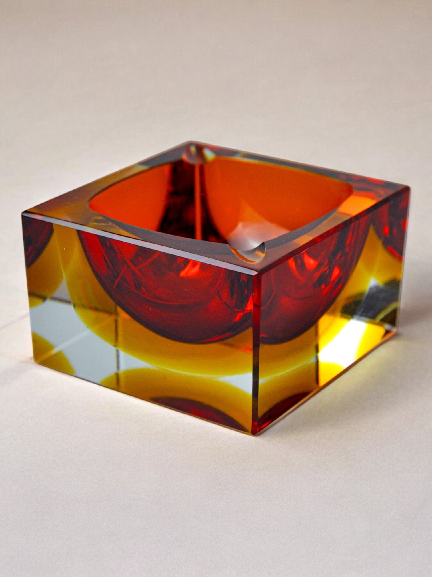 Introducing the Sommerso Ashtray by Seguso from the Murano 70s collection, available at Malamar Studio. This collectible decorative item is a square-shaped glass piece with vibrant red and yellow hues. Its smooth, glossy surface and reflective interior create stunning patterns reminiscent of the sommerso technique. It is elegantly displayed on a simple, light-colored background.