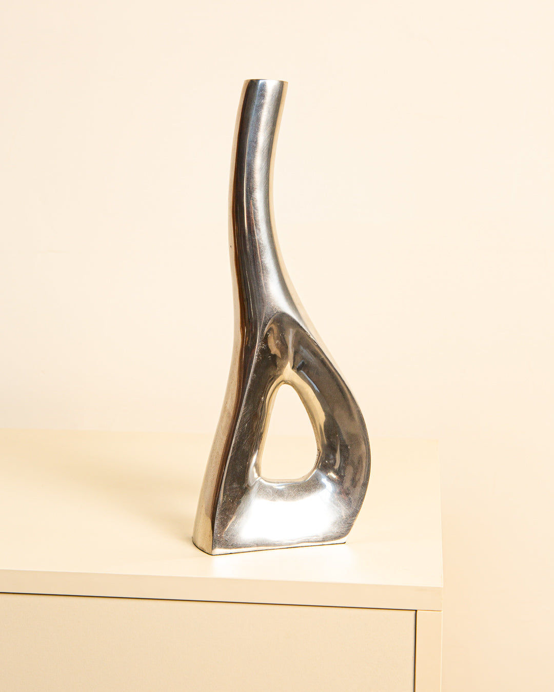 The Treaptyque Italian Vase, Aluminum 80's is a modern, abstract silver metal sculpture crafted from cast aluminum. It features a curved, elongated shape with a triangular cutout at the base and rests on a light wooden surface against a beige background for a contemporary presence.