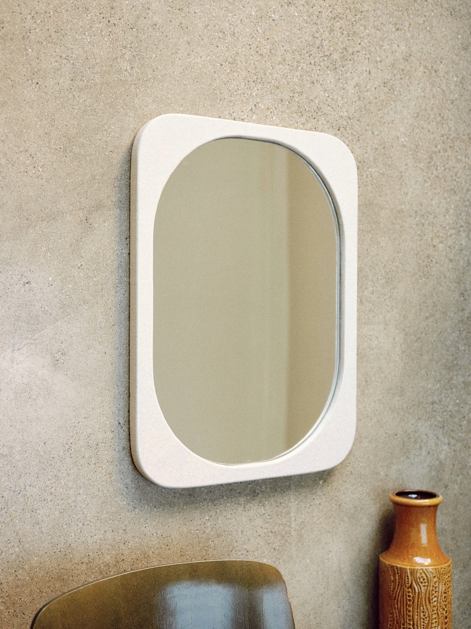 A rectangular mirror with rounded corners and a crackled enamel finish is mounted on a beige wall. The Mirror Bien Fait in Lava Stone by Bien Paris adds an artistic touch to the setting. Below it, there is a brown ceramic vase with a textured design. Part of a brown chair is visible at the bottom left of the image. The setting is minimalist and modern.
