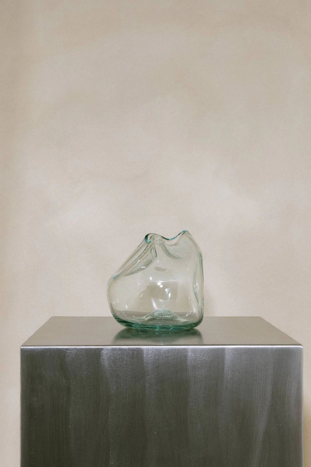 The Bubble to End all Bubbles," a hand-blown, clear recycled glass vase by Completedworks, rests elegantly on a shiny metal pedestal. A soft beige background highlights its smooth, organic form.