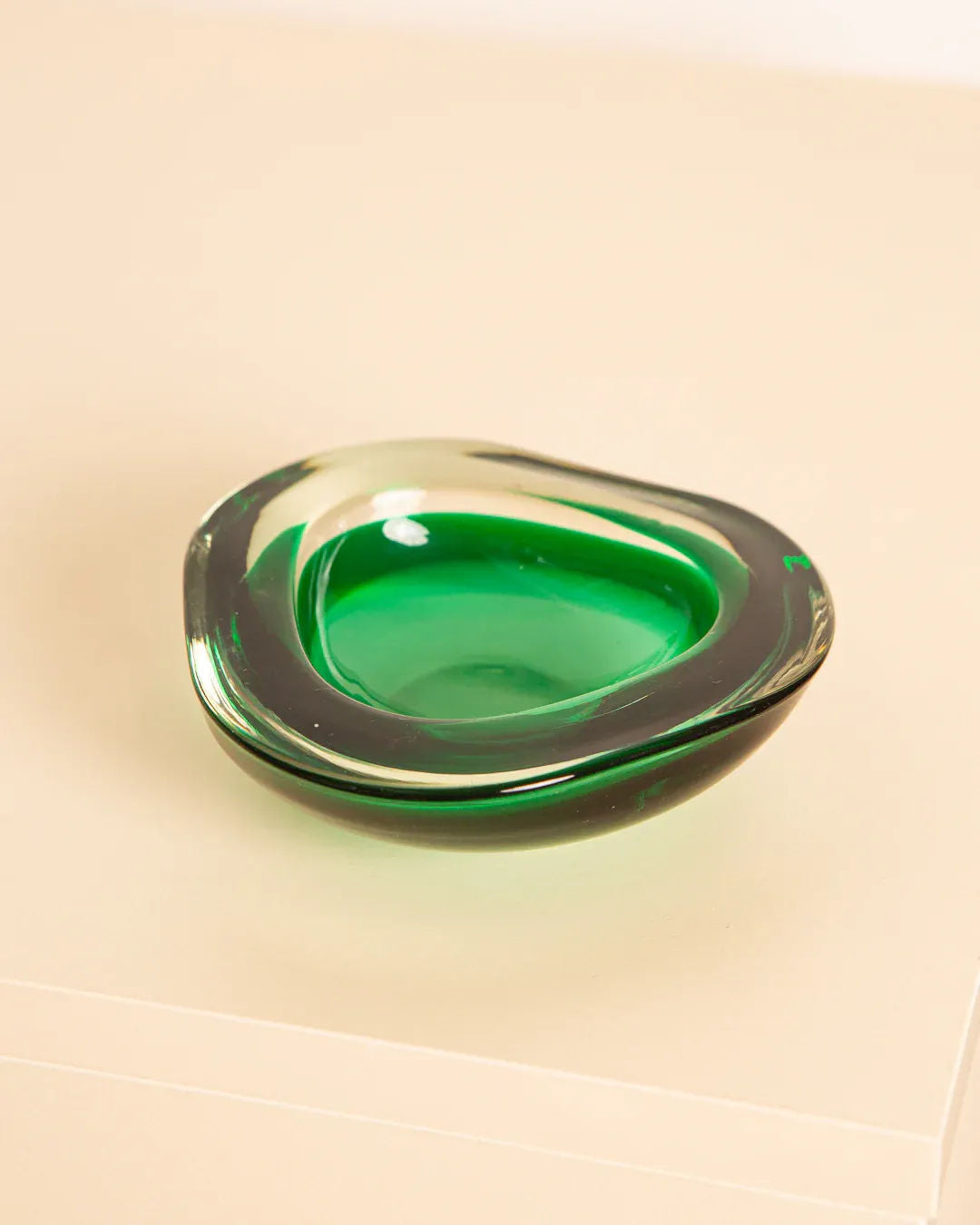 The Vide-poches "Triangle" Murano 60's by Treaptyque is a small, glossy glass bowl with an abstract, asymmetrical shape on a light beige surface. Its smooth modern design and thick clear edges reveal the green hue inside, showcasing Italian glassmaking artistry.