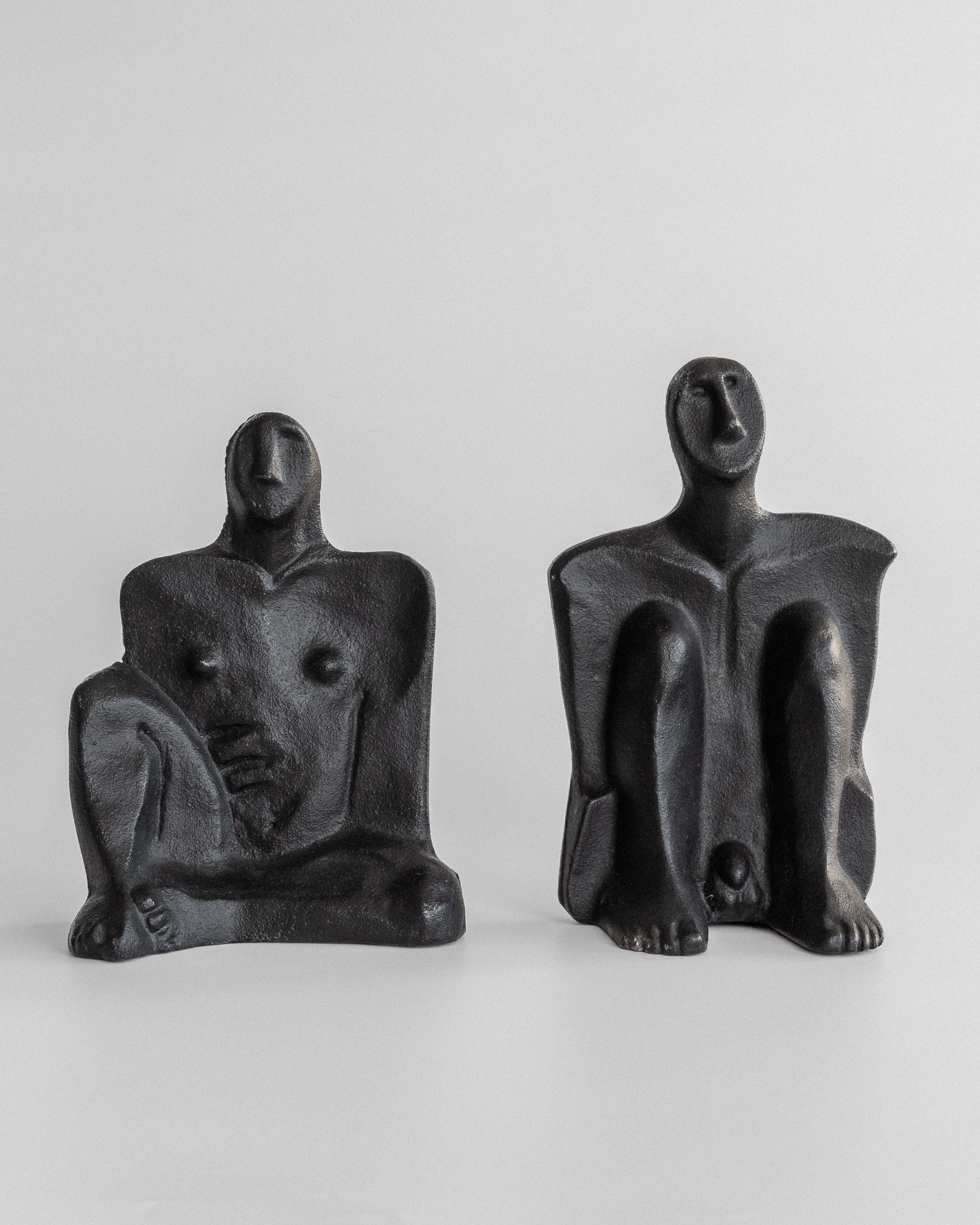 Two abstract, dark-toned cast-iron sculptures of seated figures crafted by Marita Norin for Spigel showcase stylized features and elongated limbs. These Male and Female Sculptures convey calm introspection, making them ideal contemplative bookends.
