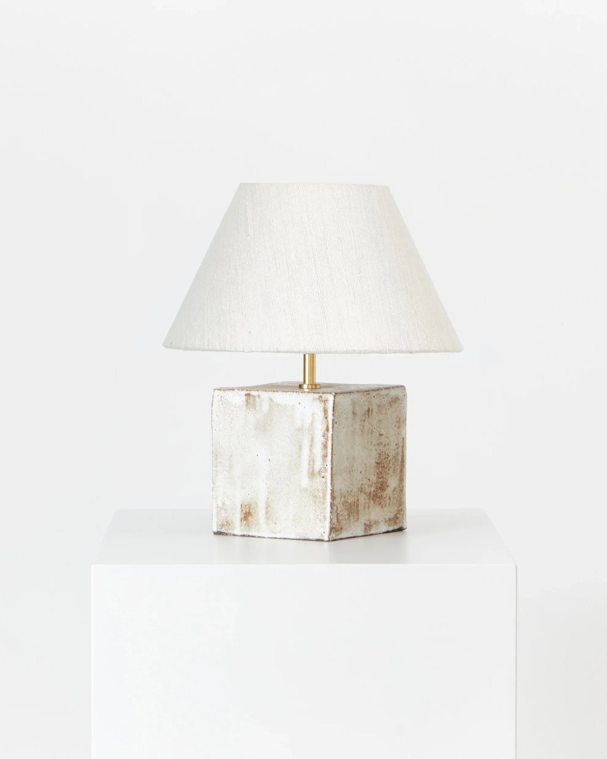 The Arouca Table Light Coral White by Project 213A, featuring a textured ceramic square base and a white fabric shade, stands on a white pedestal, highlighting the elegance of handmade lighting against a crisp white backdrop.