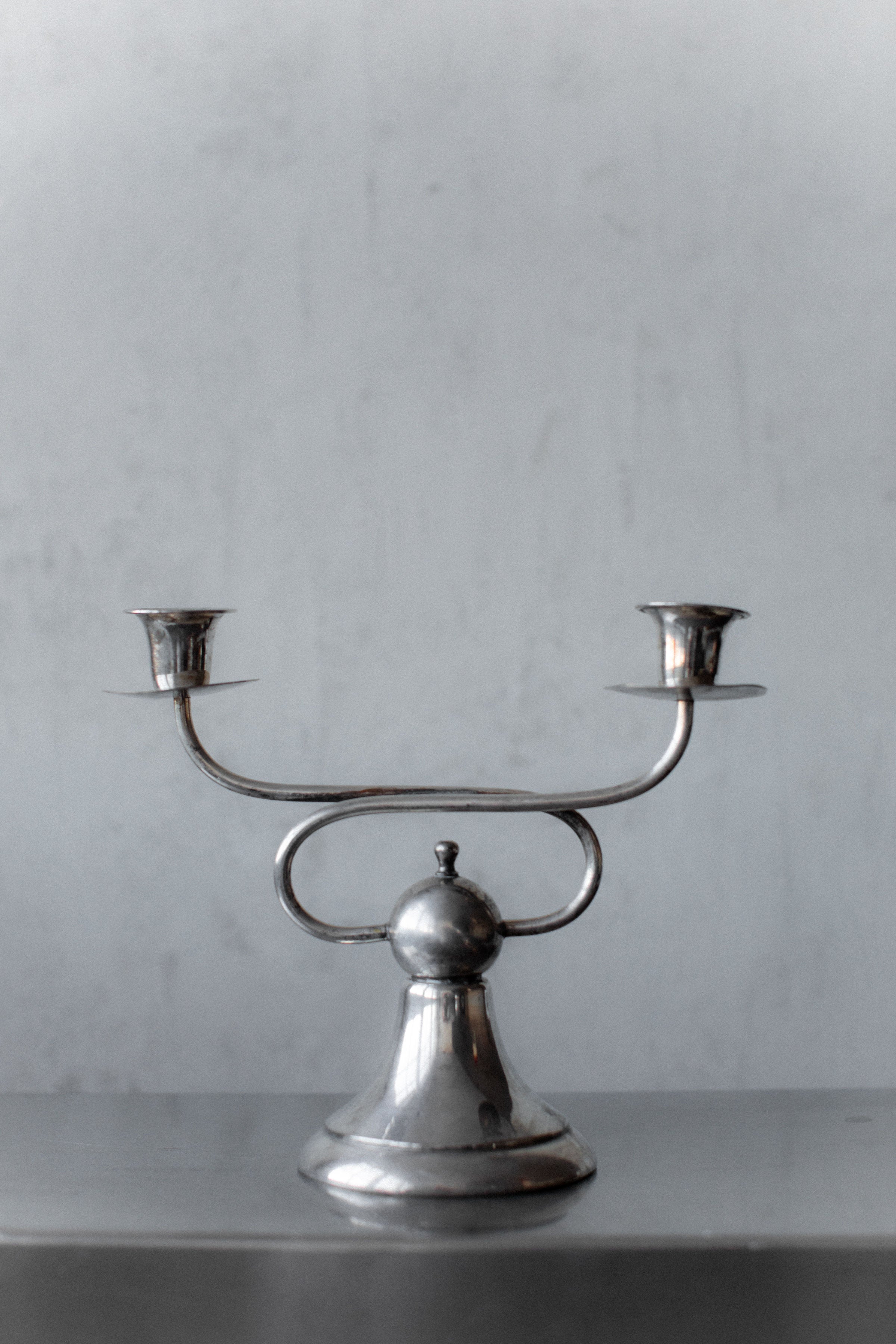 The Art Deco Candleholders by Out For Lunch exude vintage charm with two smooth, reflective arms on a gray background, featuring a central rounded base and elegant curving arms, each ending in a candle socket.