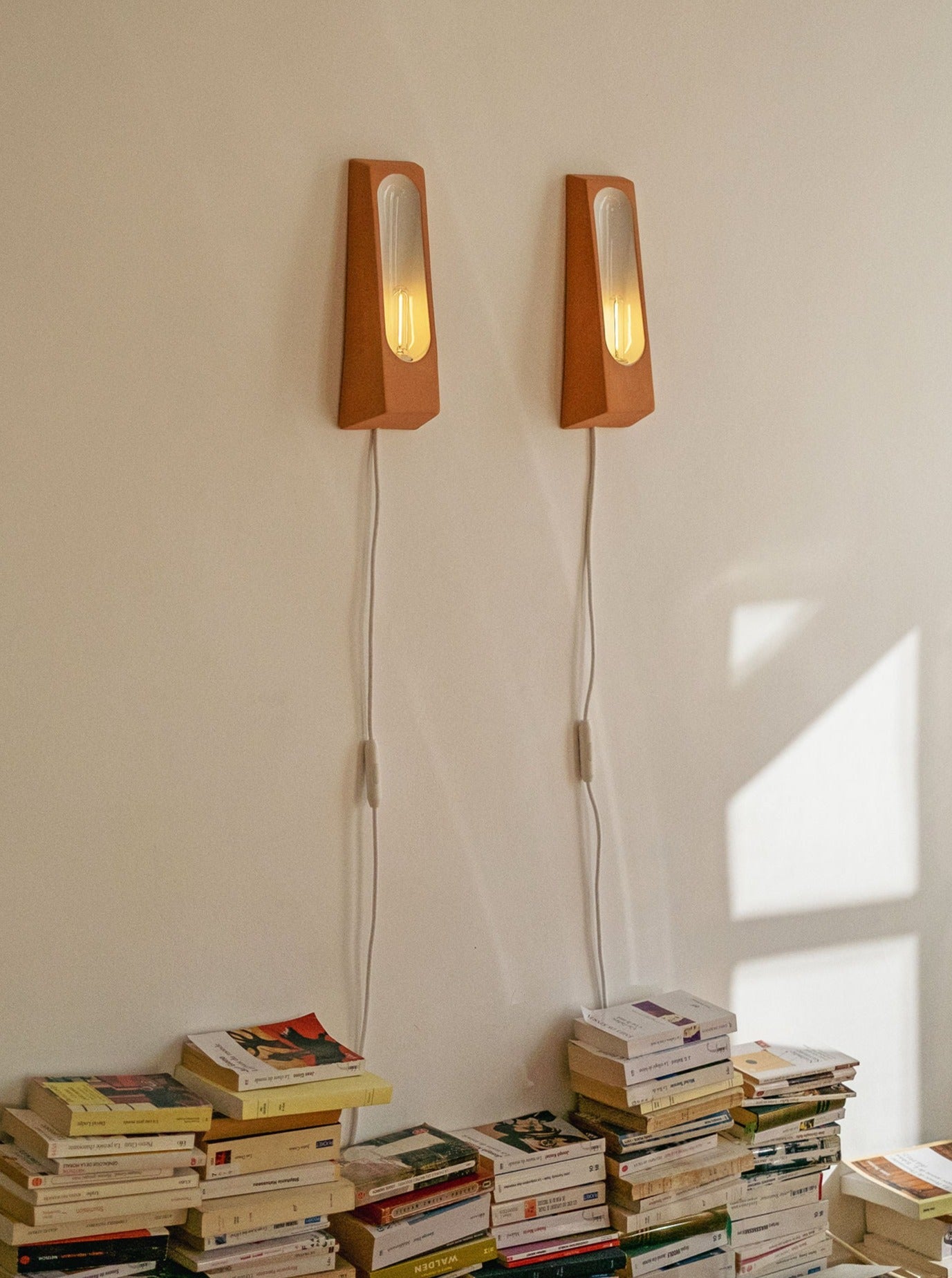 Two Alcove Terra B Mat wall-mounted ceramic enamel lamps with glowing bulbs illuminating a pile of assorted books stacked on the floor, casting soft shadows on a white wall by Violaine d'Harcourt.