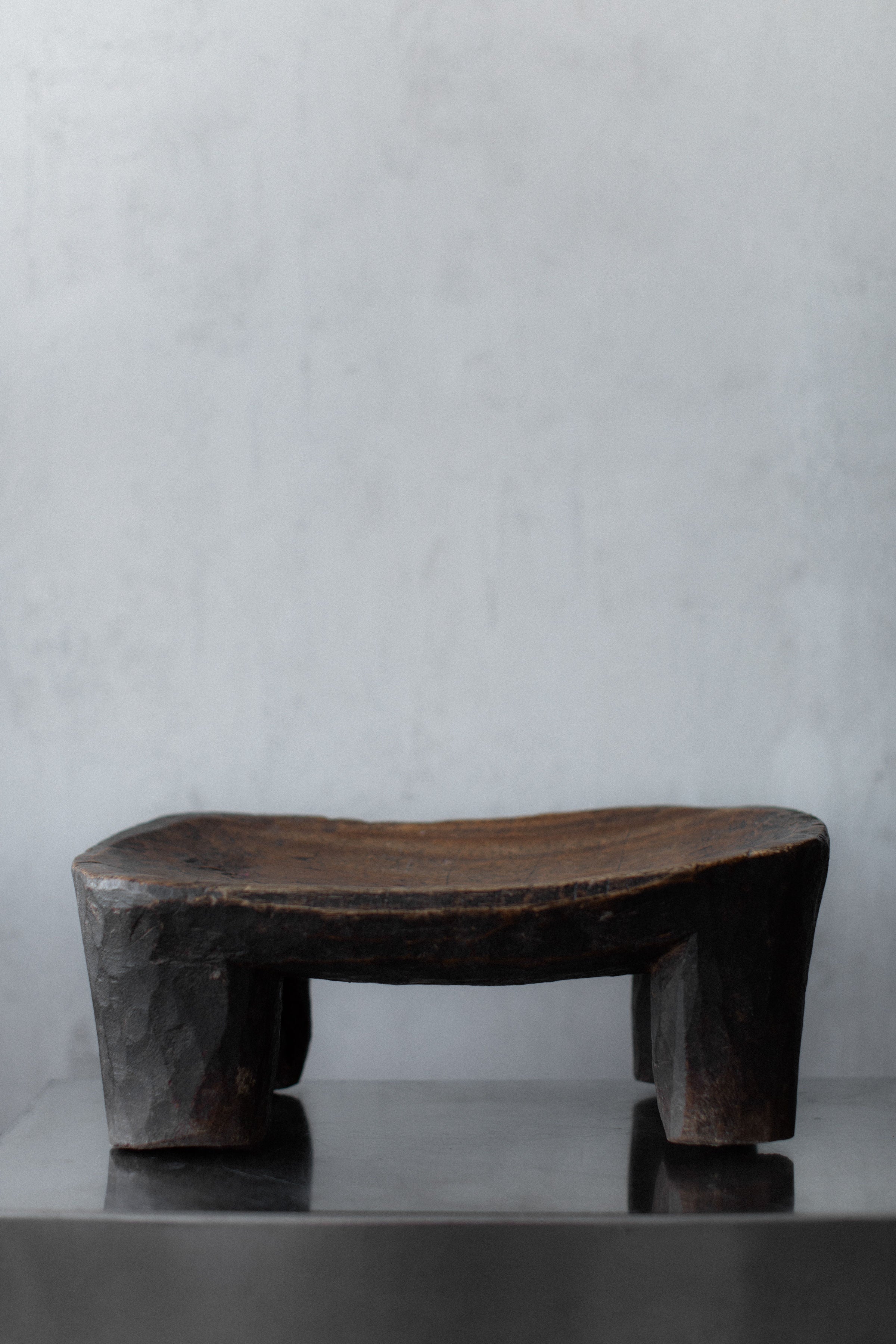 The Senufo Stool by Out For Lunch is a rustic rectangular wooden piece featuring a minimalist design with a curved top and four thick legs, set against a plain gray background. Its weathered look highlights the natural wood grain, embracing global design aesthetics.