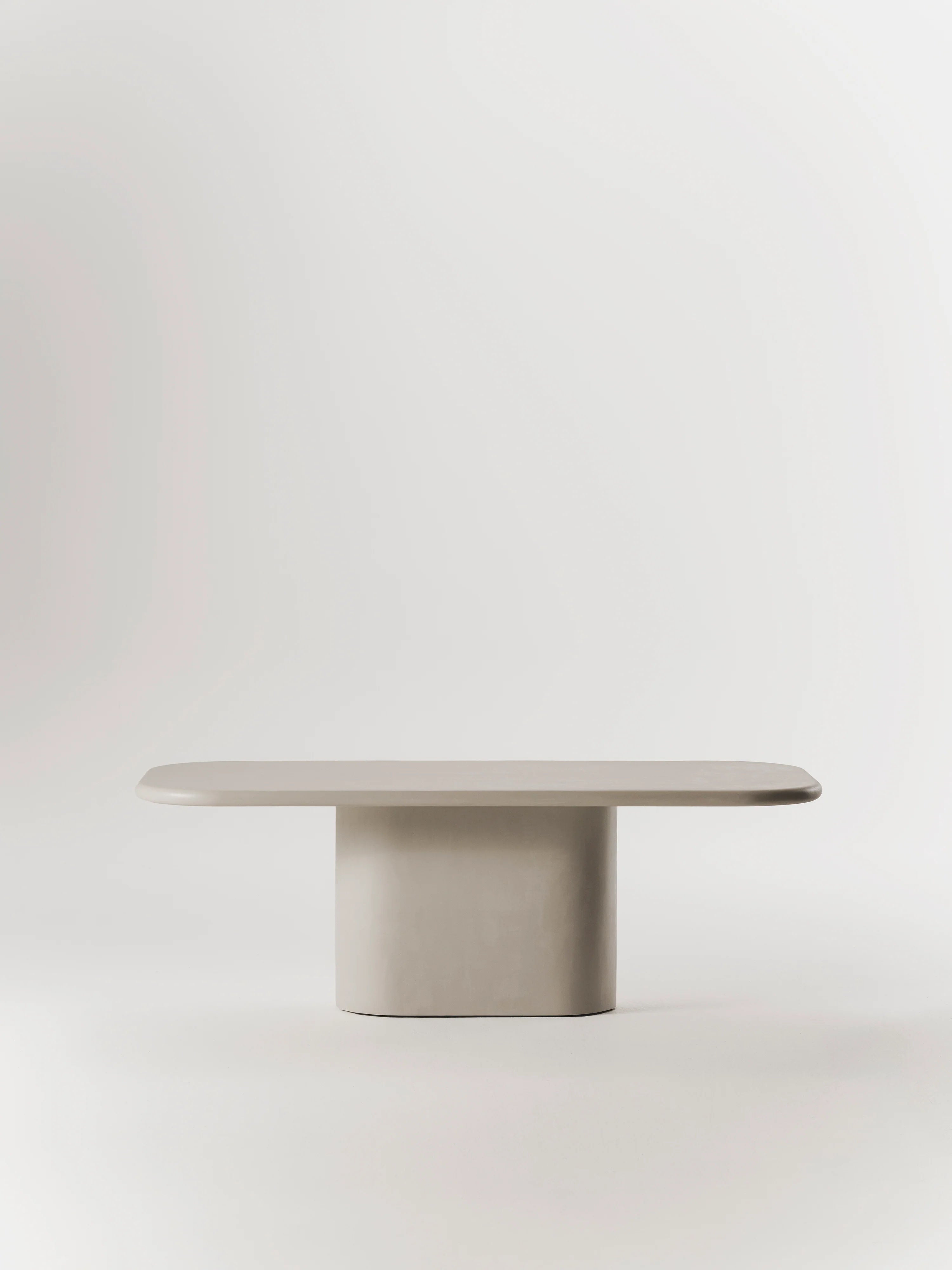 The MarlotBaus Lucca Dining Table, a modern minimalist piece with rounded edges, rests against a white background. Its smooth micro cement top and sturdy oval central leg blend elegance and durability effortlessly.