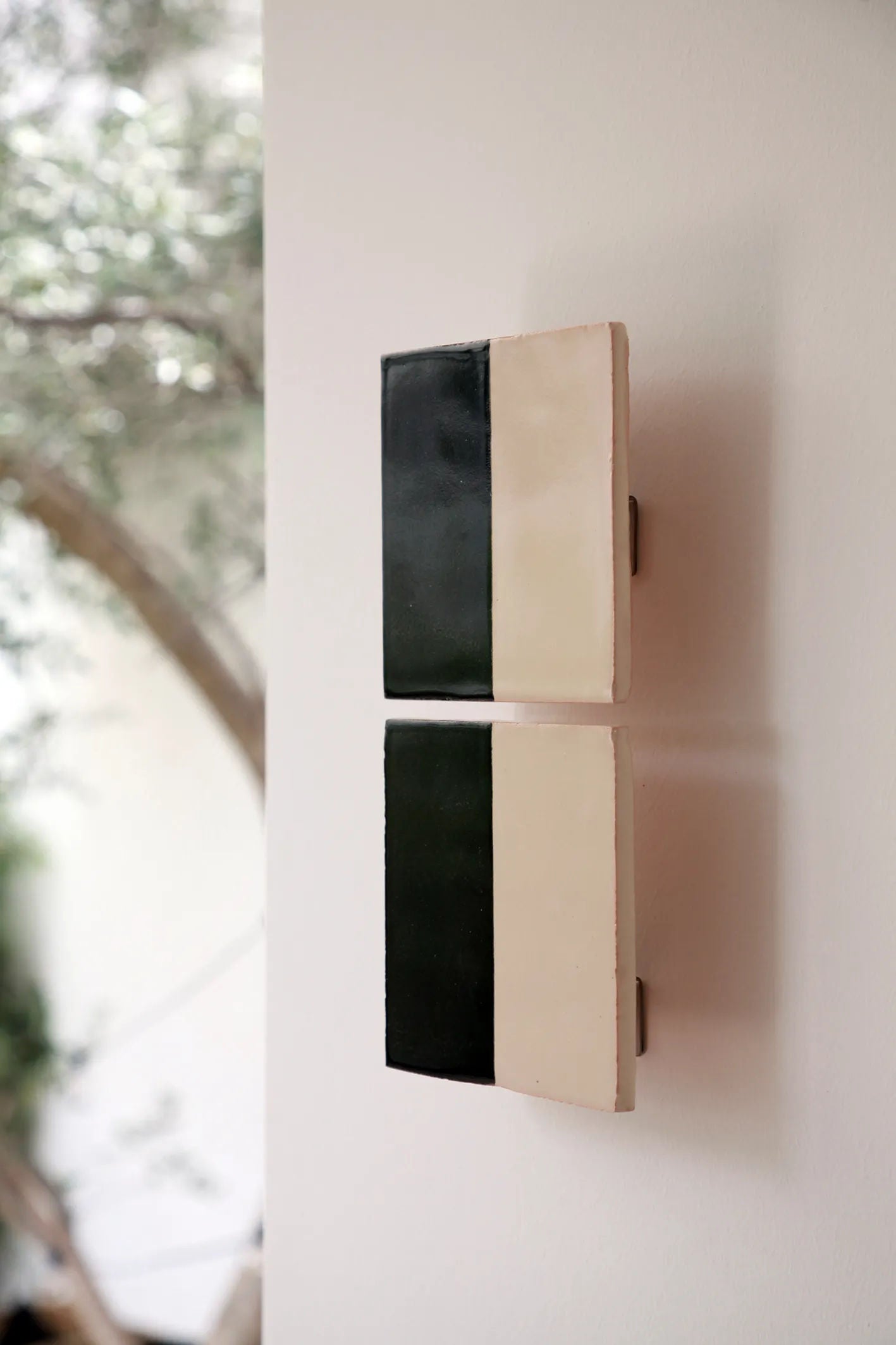 Two rectangular Tiles Line N by Violaine d'Harcourt, divided vertically into black on the left and cream on the right, are mounted on a white wall. Blurred greenery in the background hints at an outdoor setting.