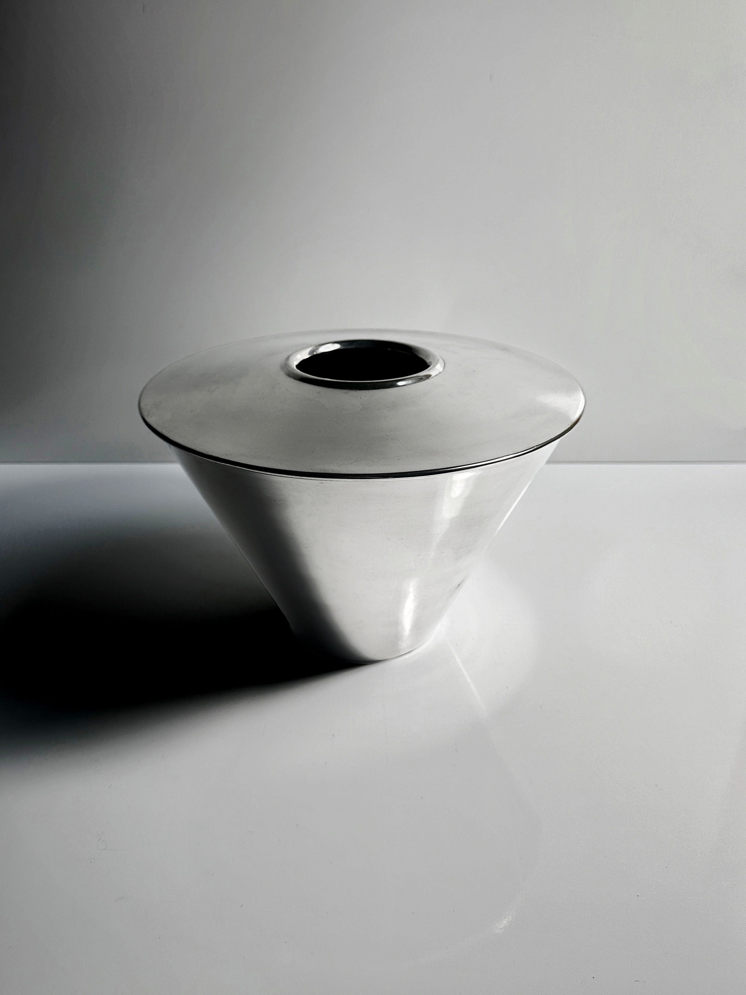 A sleek, polished metal cone-shaped Heavy Silver-Plated Vase from Fink Germany by Dodo Vintage features a circular opening on top. Set against a minimalist gray and white background, the lighting casts subtle shadows highlighting its smooth surface.