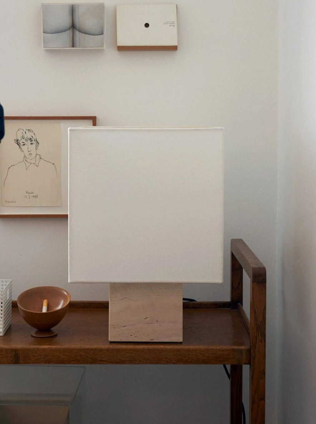 A minimalist, cozy corner featuring a Roma I Squared White Travertine lamp by Nassi Lamps on a wooden side table. Artwork hangs on the wall above; one piece is a monochrome photograph, another a modernistic abstract, and a simple pencil sketch. A small bowl and a candle are also on the table.