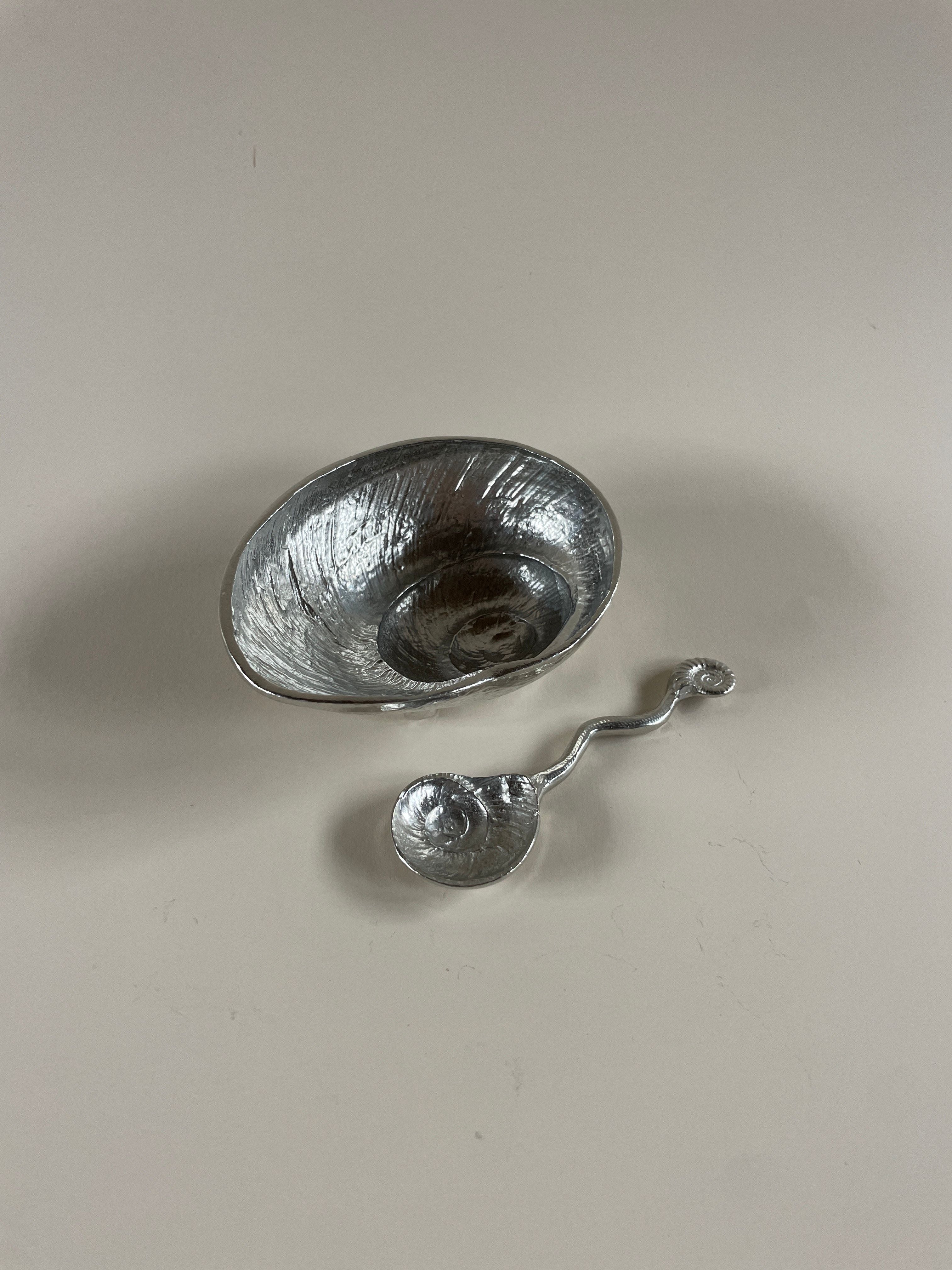 The Shell Dish & Spoon by Maud Vaughan, featuring a small, textured vintage polished pewter dish with a wavy design, sits beside its matching spoon on a plain light background.