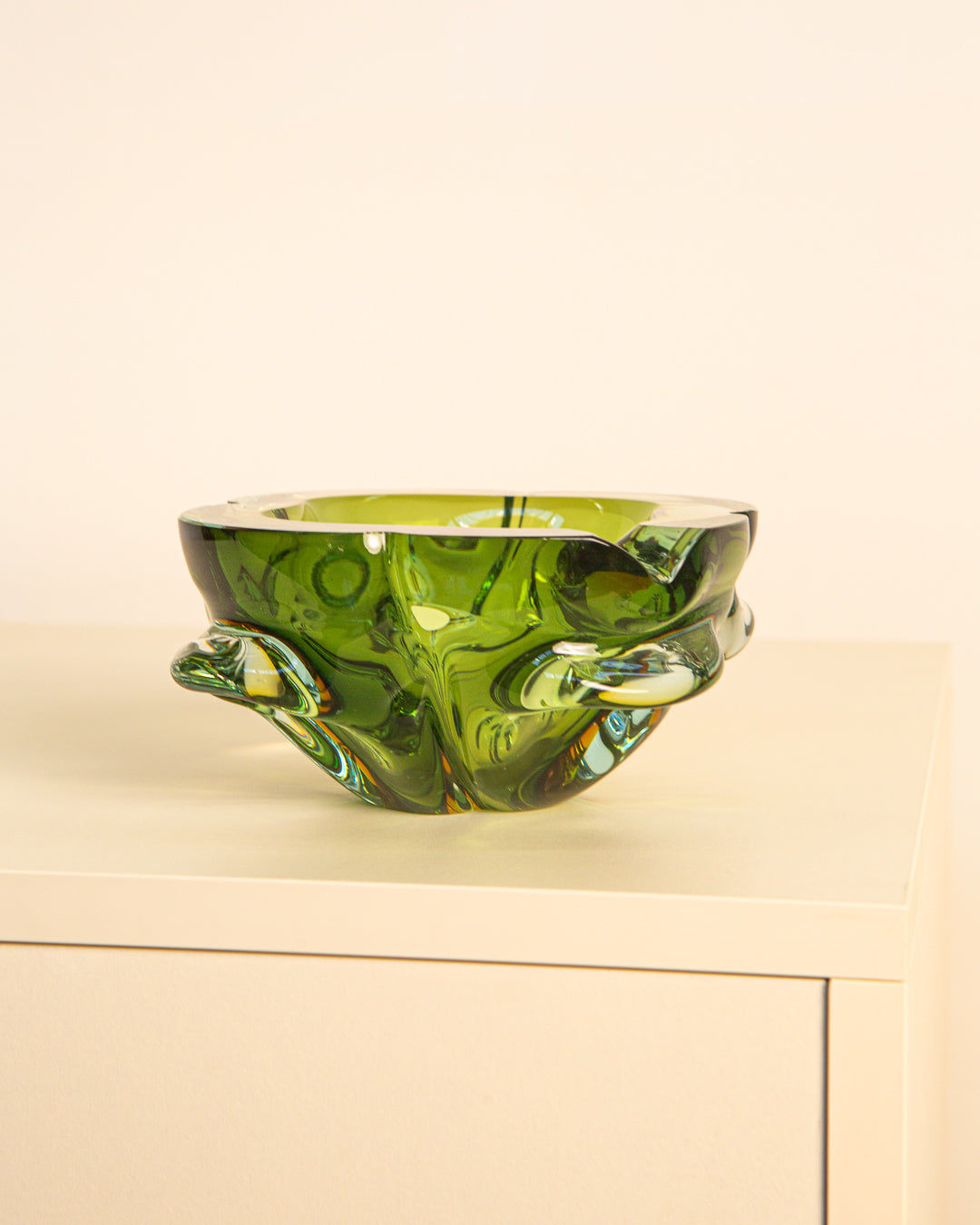 The Treaptyque Dark Green Murano Glass “Fleur” Ashtray from the 60s, with a wavy abstract design, sits on a light cabinet against a beige backdrop, embodying iconic Italian style.