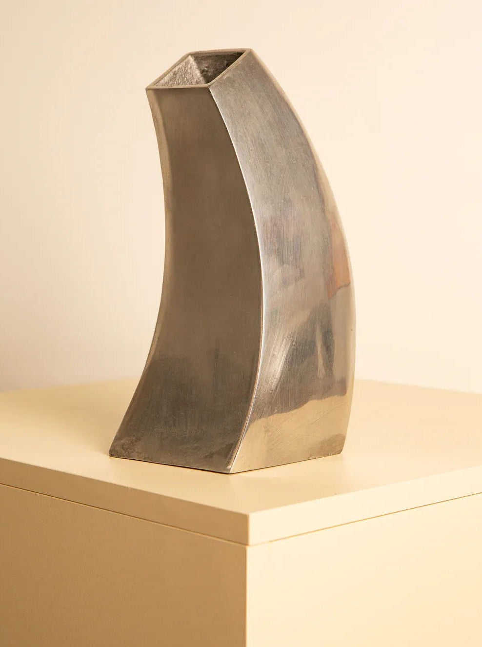 A sleek, modern silver *80s Metal Vase Sculpture* by Treaptyque with a curved, asymmetrical design reminiscent of '80s metal sculpture stands on a beige surface against a light background. The vase has a shiny finish and an angled, rectangular opening at the top.
