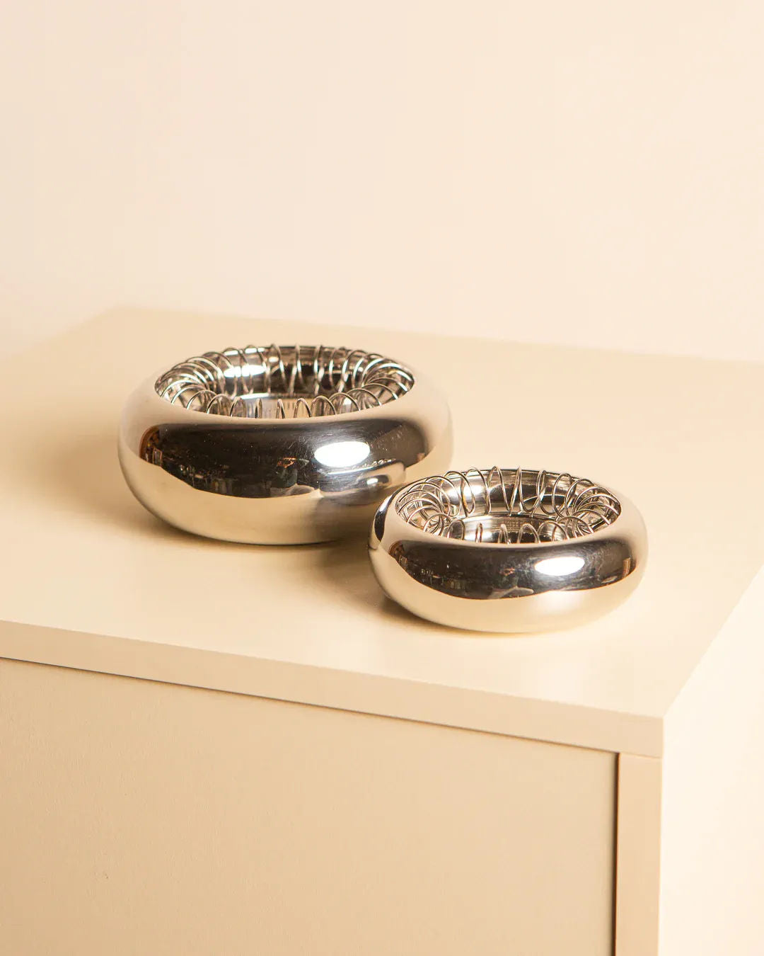 Two shiny silver bowls, crafted by Treaptyque, echo Italian design with textured interiors, resting on a cream-colored surface. They differ in size, with the larger on the left. The neutral beige background evokes Achille Castiglioni's "Spirale" Ashtrays for Alessi 90s.
