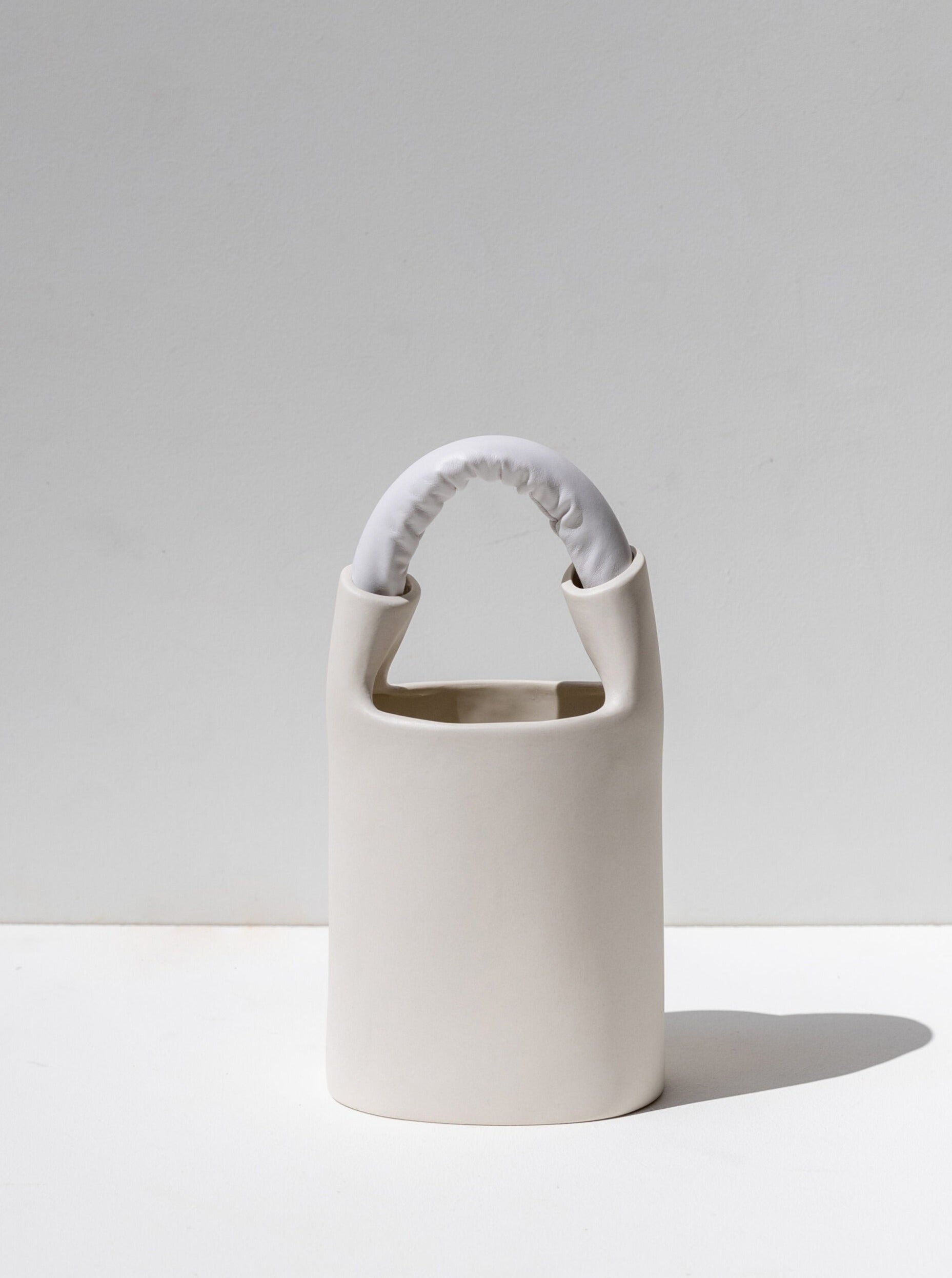 The Bag Vase Apparel by Lisa Allegra is a minimalist ceramic vase featuring a smooth cylindrical body with a handle at the top, partially wrapped in monochrome fabric-like material. This sculptural object vase is displayed against a light gray background, casting a small shadow to its right.