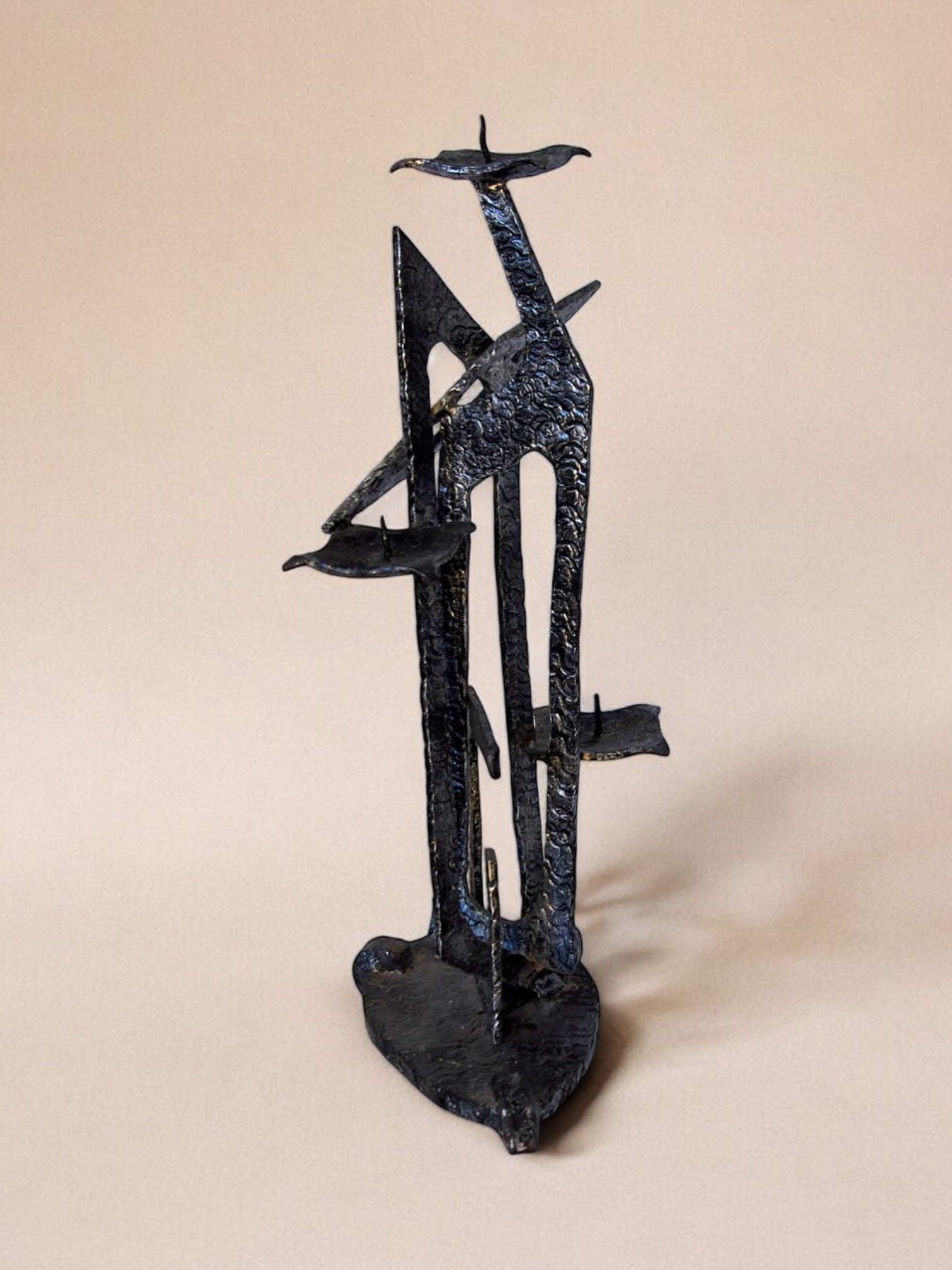 The Malamar Studio Brutalist candle holder, inspired by David Palombo's 1960s design, features a metal sculpture with a textured dark finish. It resembles a stylized bird with outstretched wings and stands on a triangular base against a neutral background.