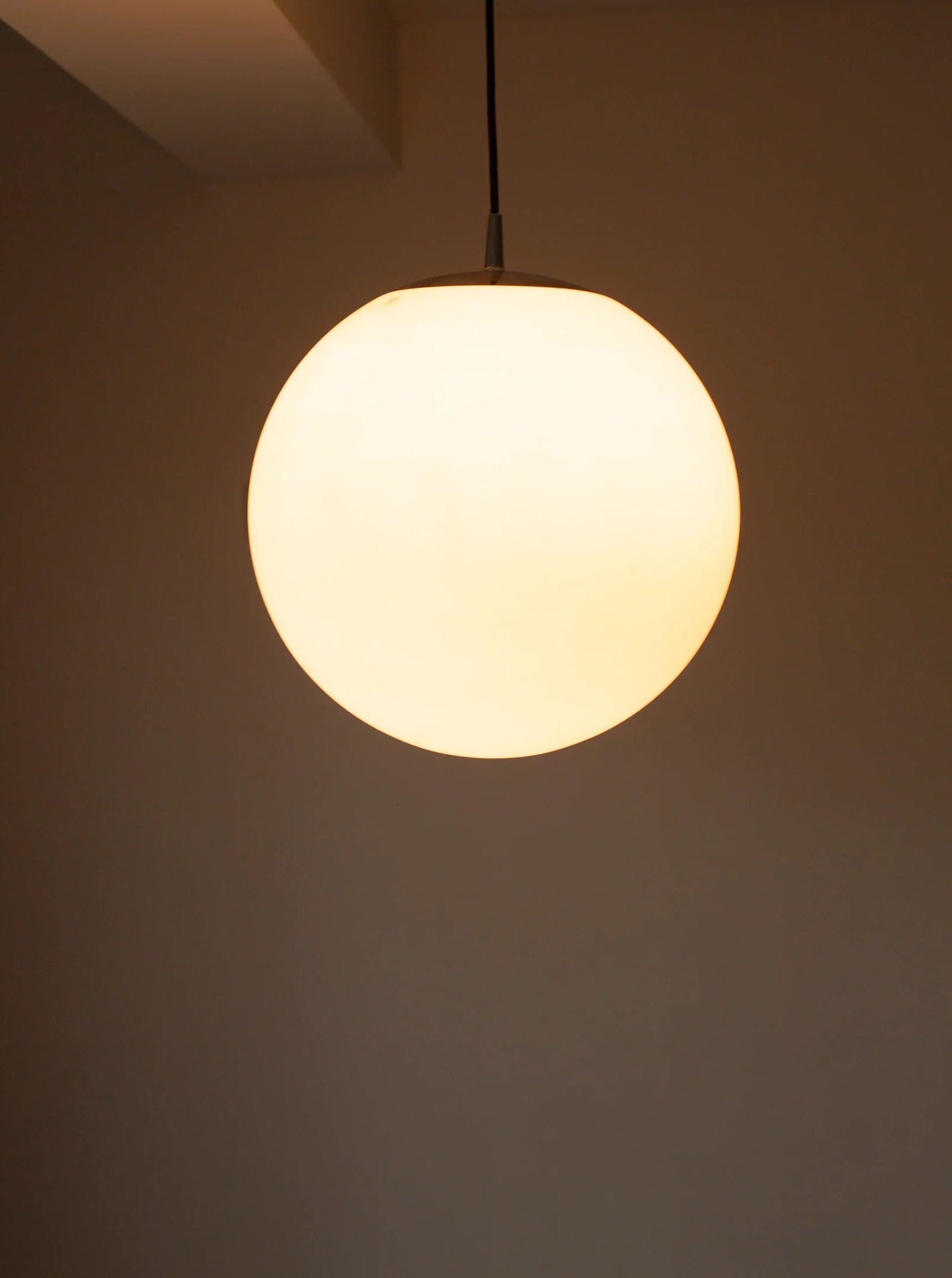 A Glass Globe Lamp Peill & Putzler emits a soft, warm glow against a plain, beige background. The light is suspended from a thin, dark cord, casting subtle shadows and creating a minimalist, serene ambiance. This fixture is reminiscent of vintage CU-RA-TED designs.