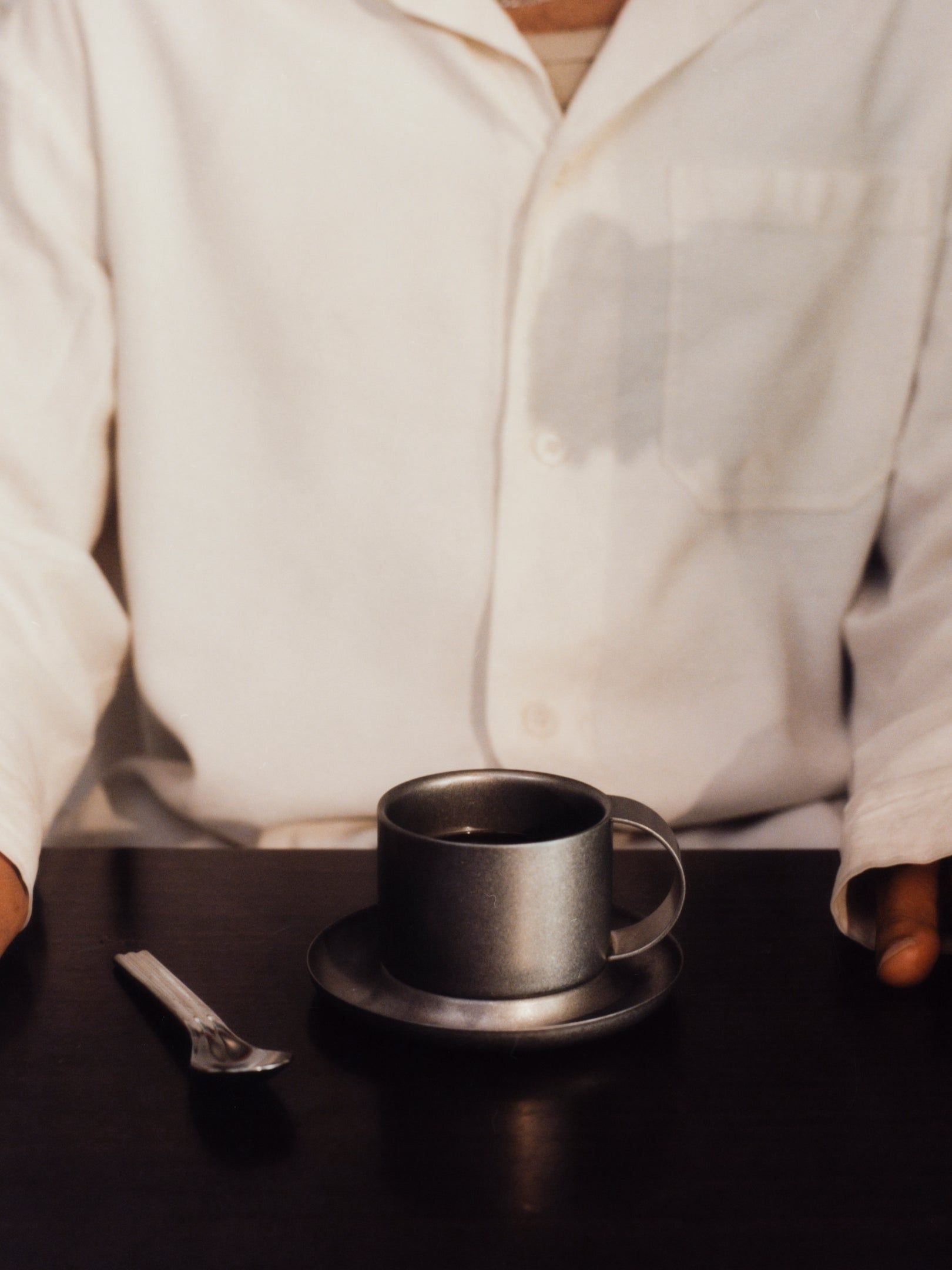 A person in a white shirt with a coffee stain sits at a dark table with Service Projects' The Arlo 03 Cup and saucer, made of stonewashed stainless steel, filled with coffee, and a spoon next to the cup.