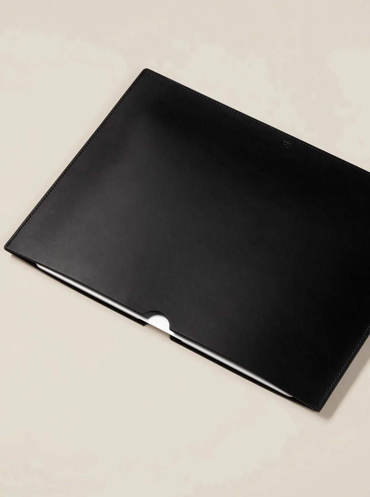A stylish black Paradise Row Leather Laptop Sleeve, made from ethically sourced materials, lies on a neutral surface. Its minimalist design includes a small semicircular opening in the center, offering a glimpse of the laptop inside.