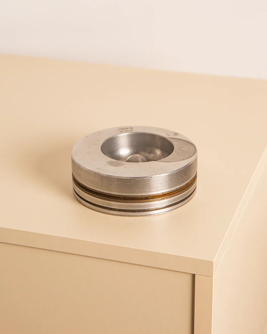 The Italian "Piston" ashtray in 80's metal by Treaptyque exudes vintage charm with its round, metallic design and features a transparent dome filled with small stones. It sits on a beige, minimalist desk against a light background, adding an elegant touch reminiscent of Milanese design.